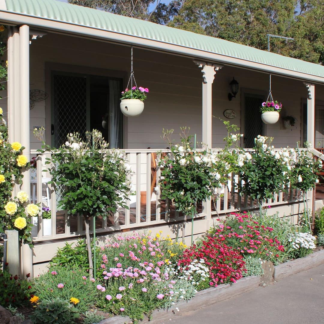 Charming cottage with beige siding and light green roof, a covered porch, and a lush garden surrounding the home.