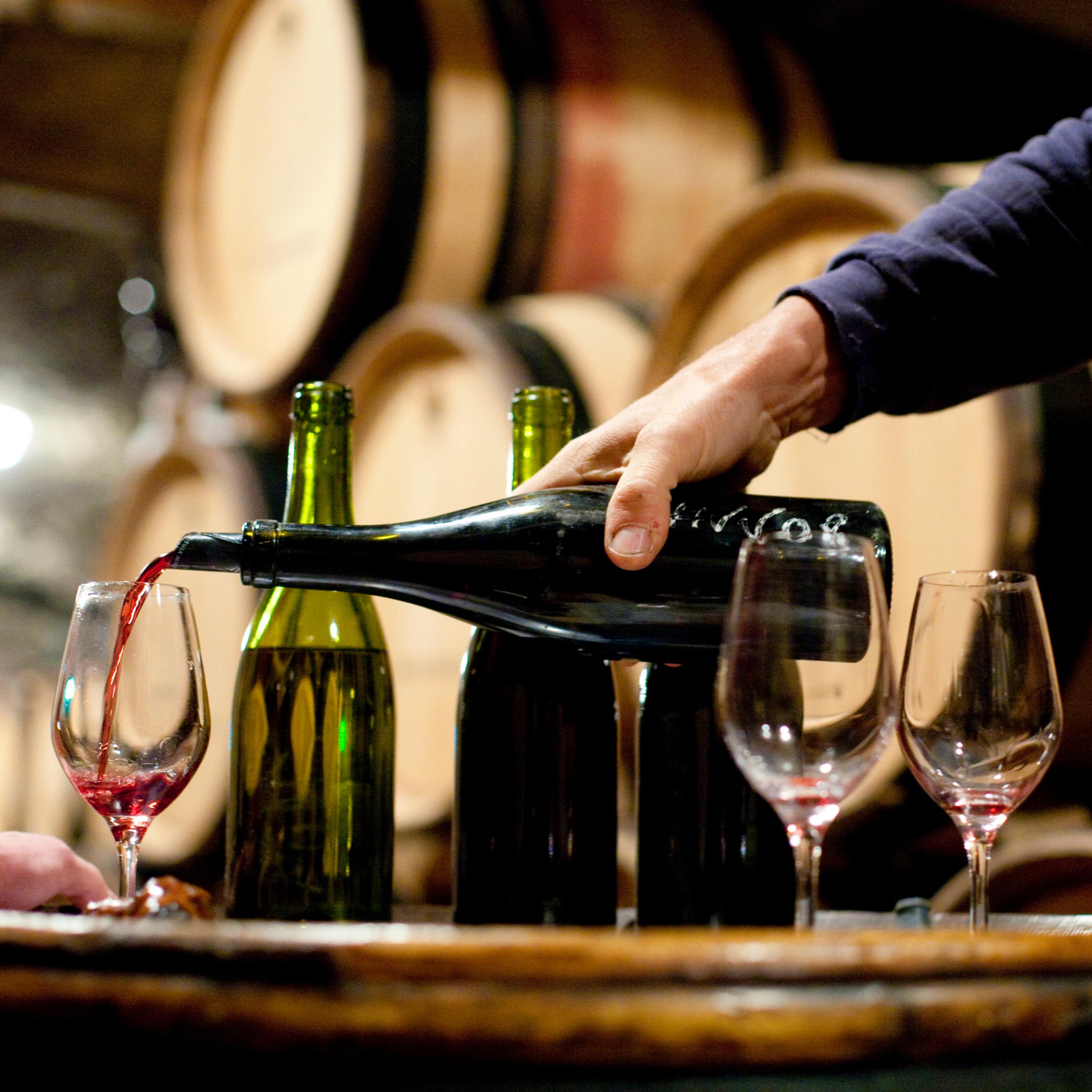 Red wine being poured for a tasting with barrels of wine in the background.