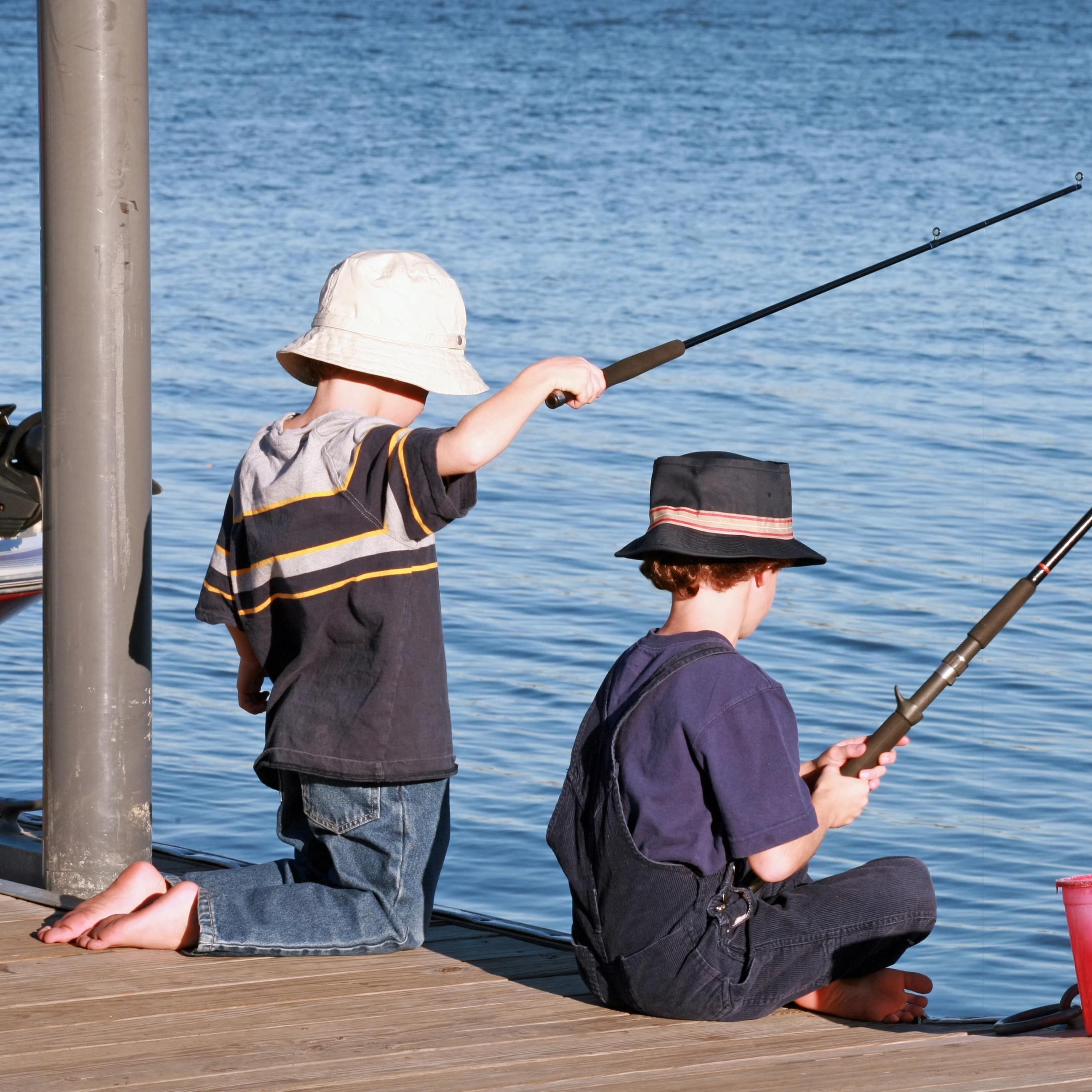 Two young boys with fishing hats on fishing off dock with red bucket waiting