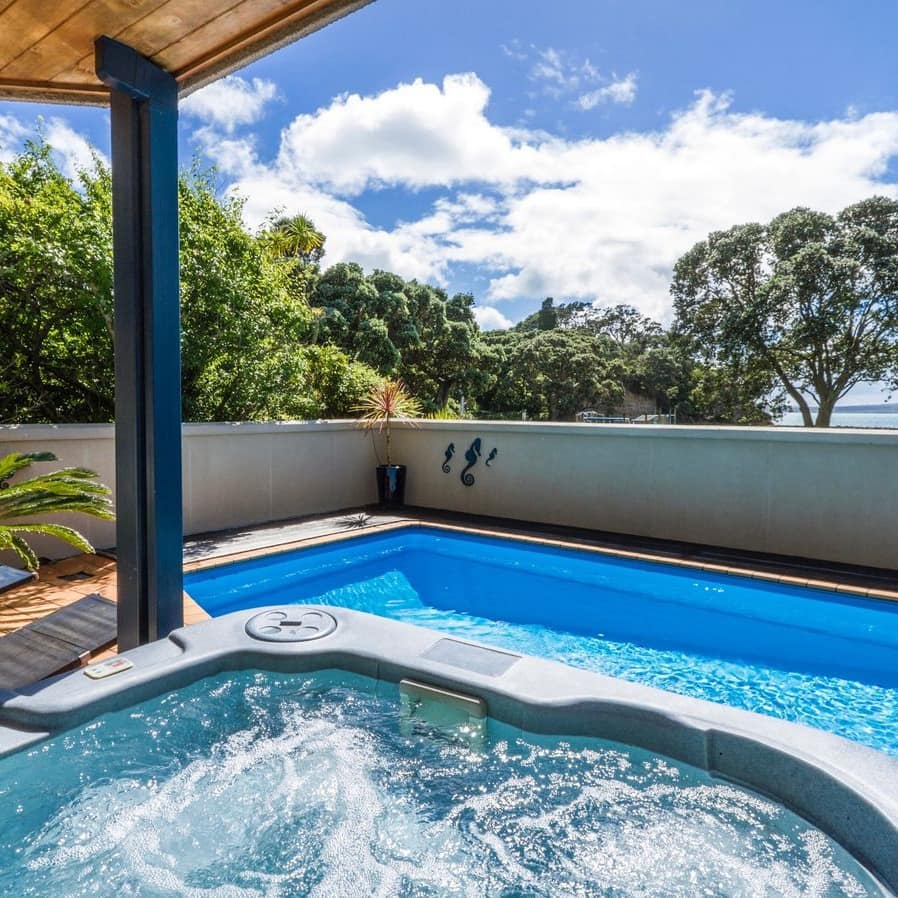 The edge of a hot tub and a pool in the backyard of a holiday home with a view of the ocean in the distance