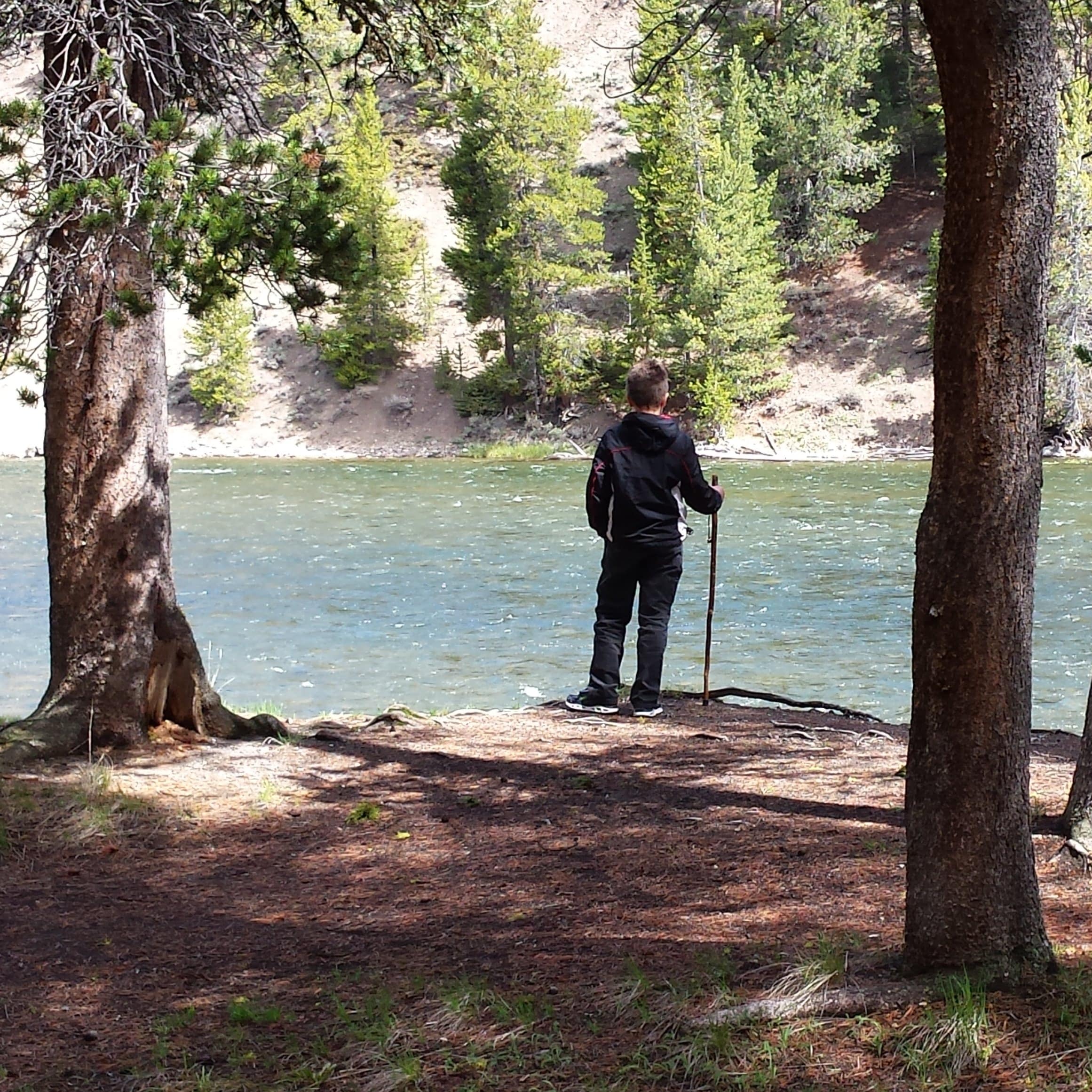 A young boy standing on the bank of a lake surrounded by trees.