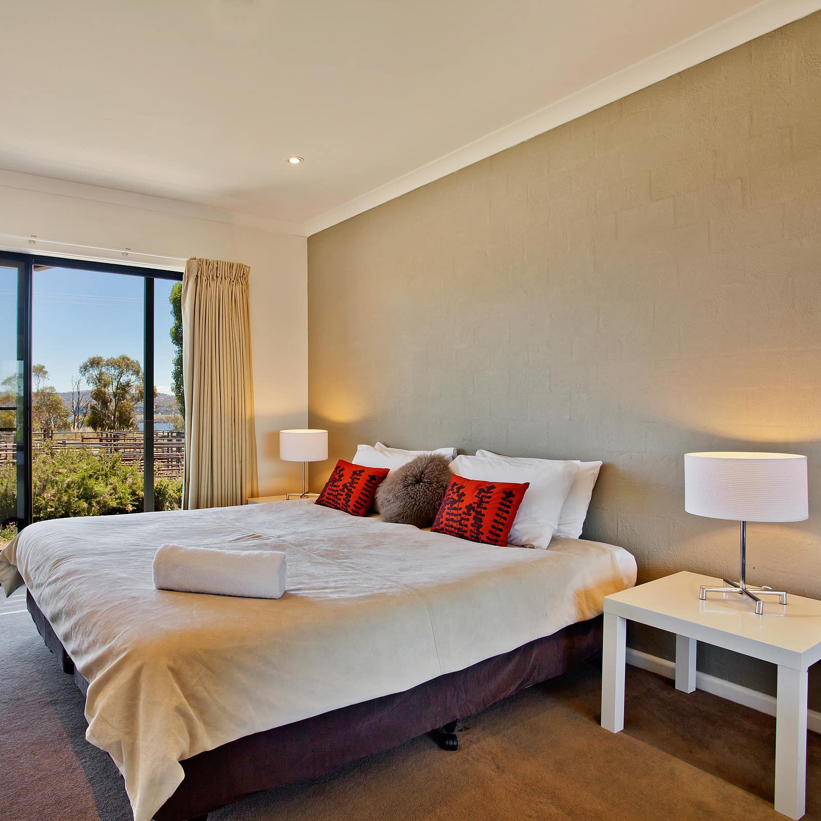 Spacious bedroom, king size bed, white linen, red pillows, tan accent wall, grey carpet, and tall windows with lake views.