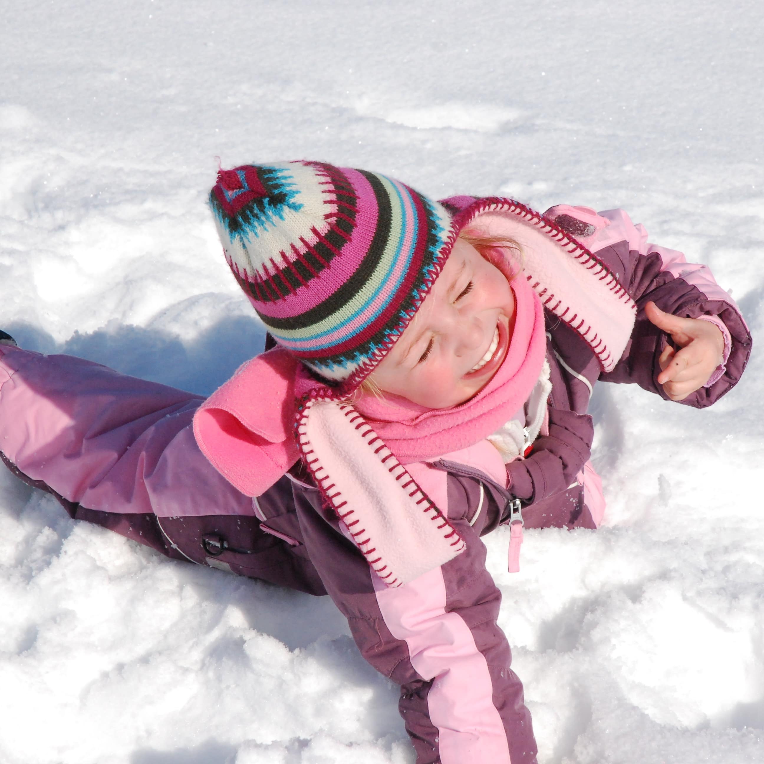 Cute little girl lying in the snow with pink snowsuit with thumb up and silly face