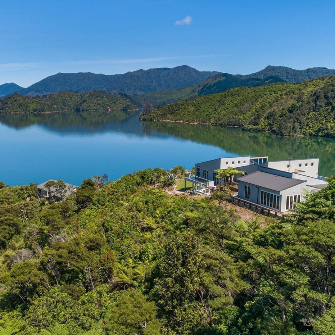 Aerial view of a large family home nestled among green trees on clear blue waters in South Island