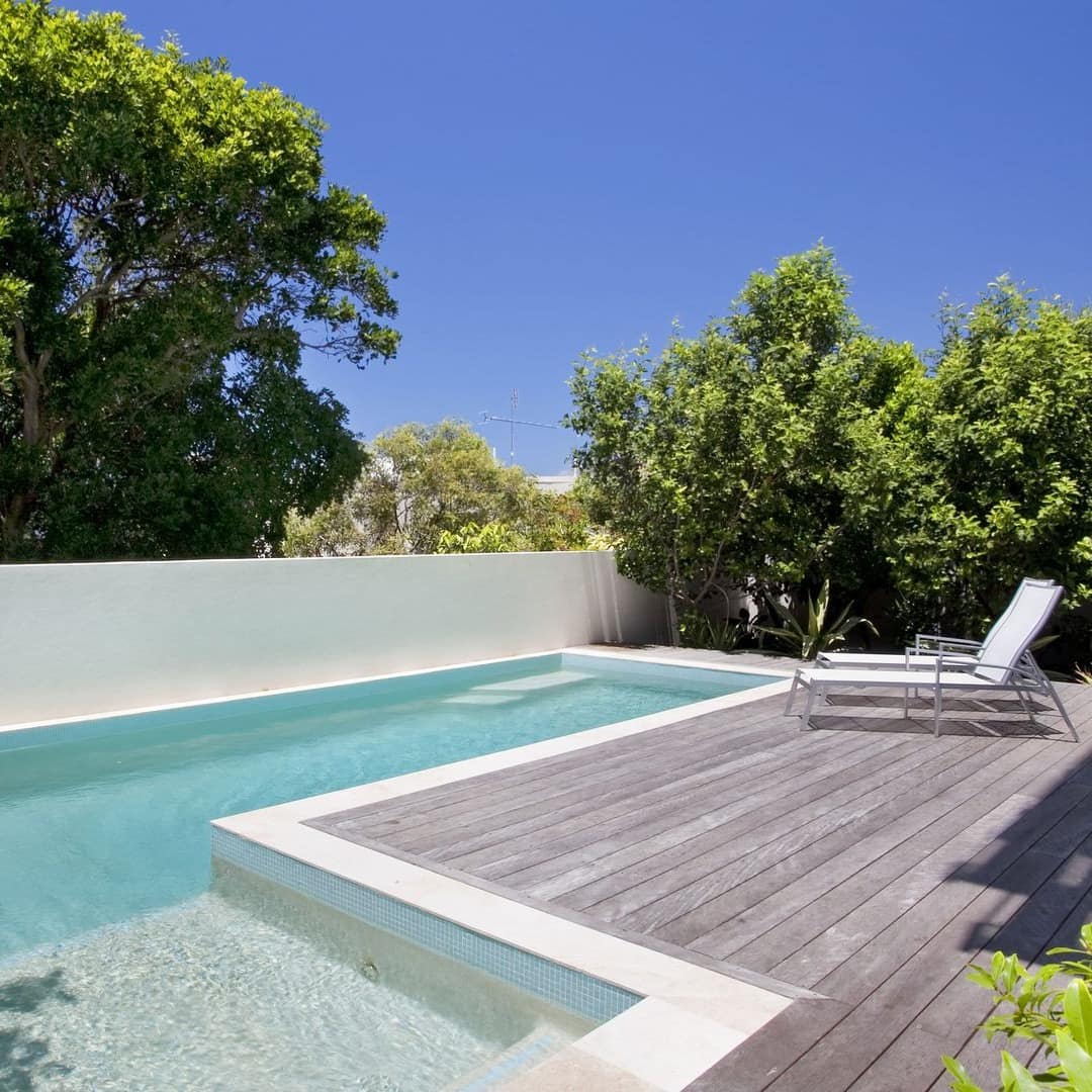 View of an inground pool with wooden pool deck, white lounge chairs, surrounded by lush vegetation.
