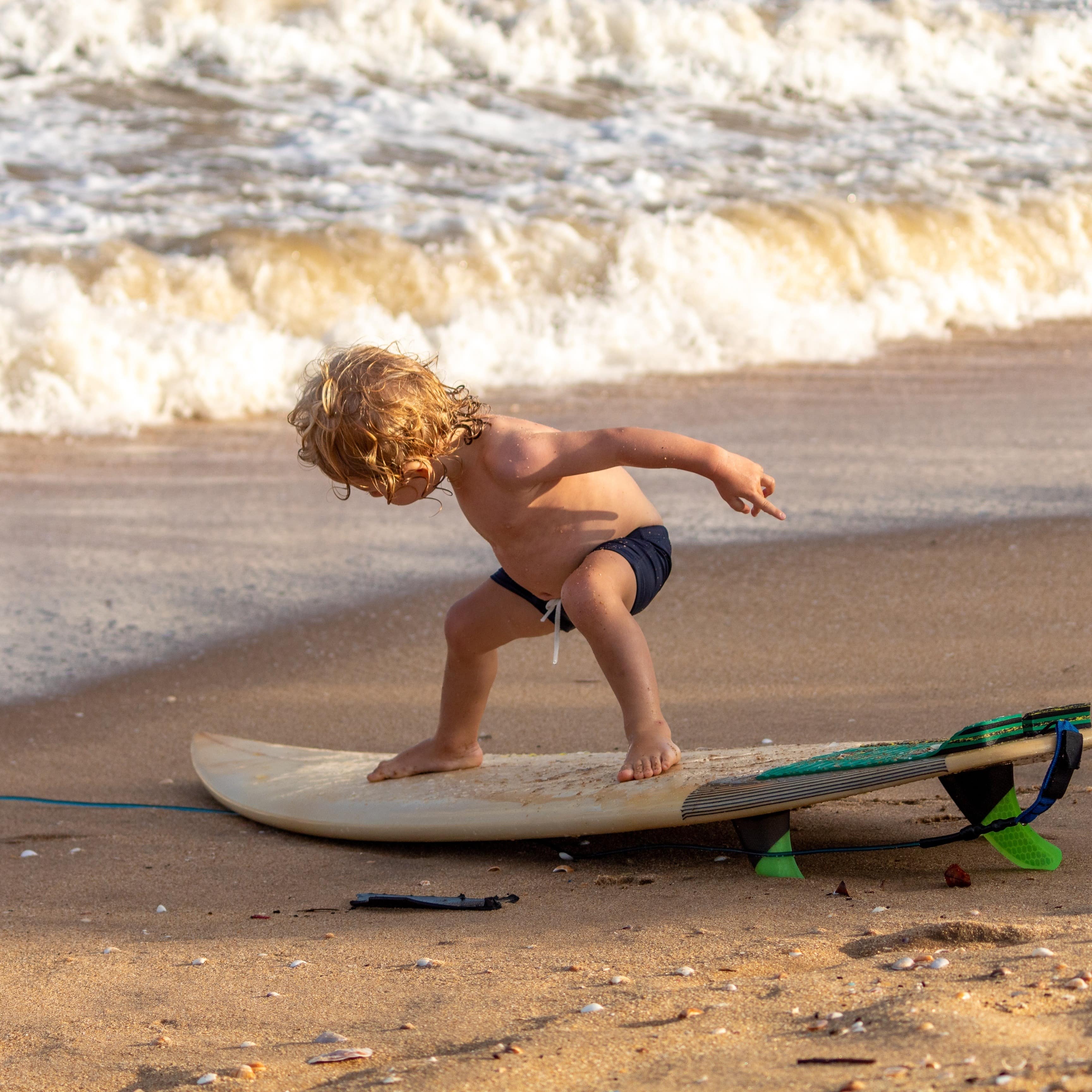 Toddler boy at on surfboard at surf’s edge with waves in background