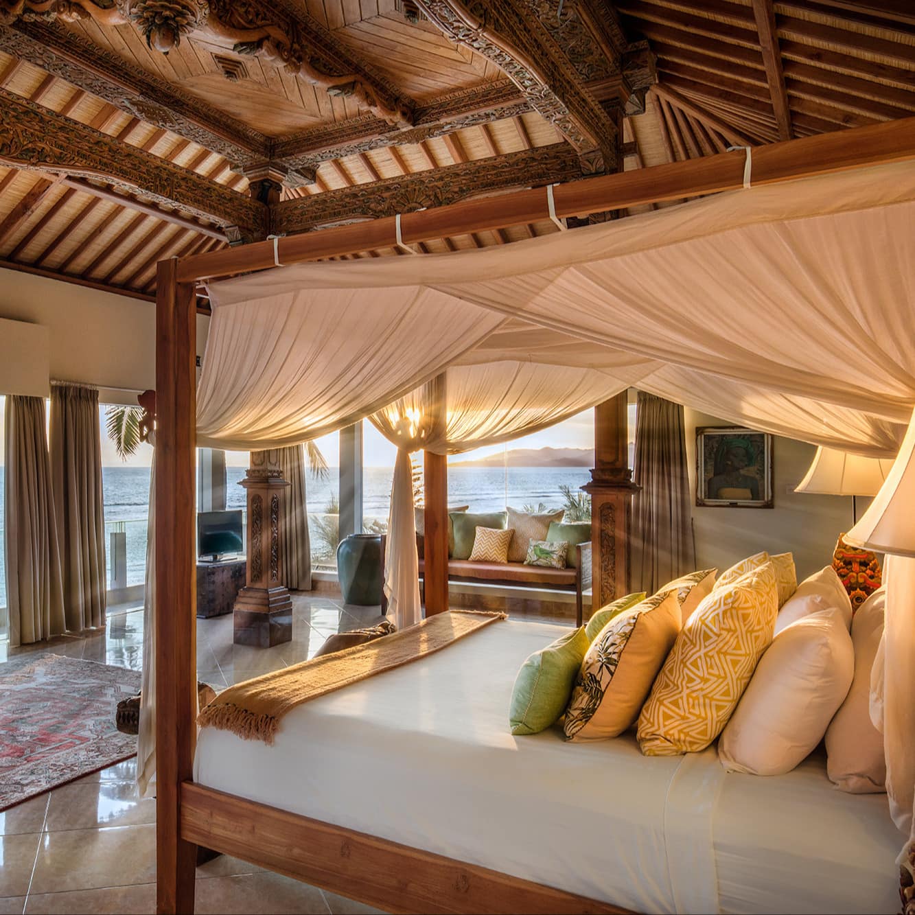 Sunny bedroom with a king size canopy bed, ornately carved wood ceiling, sliding glass doors to a balcony with ocean views.