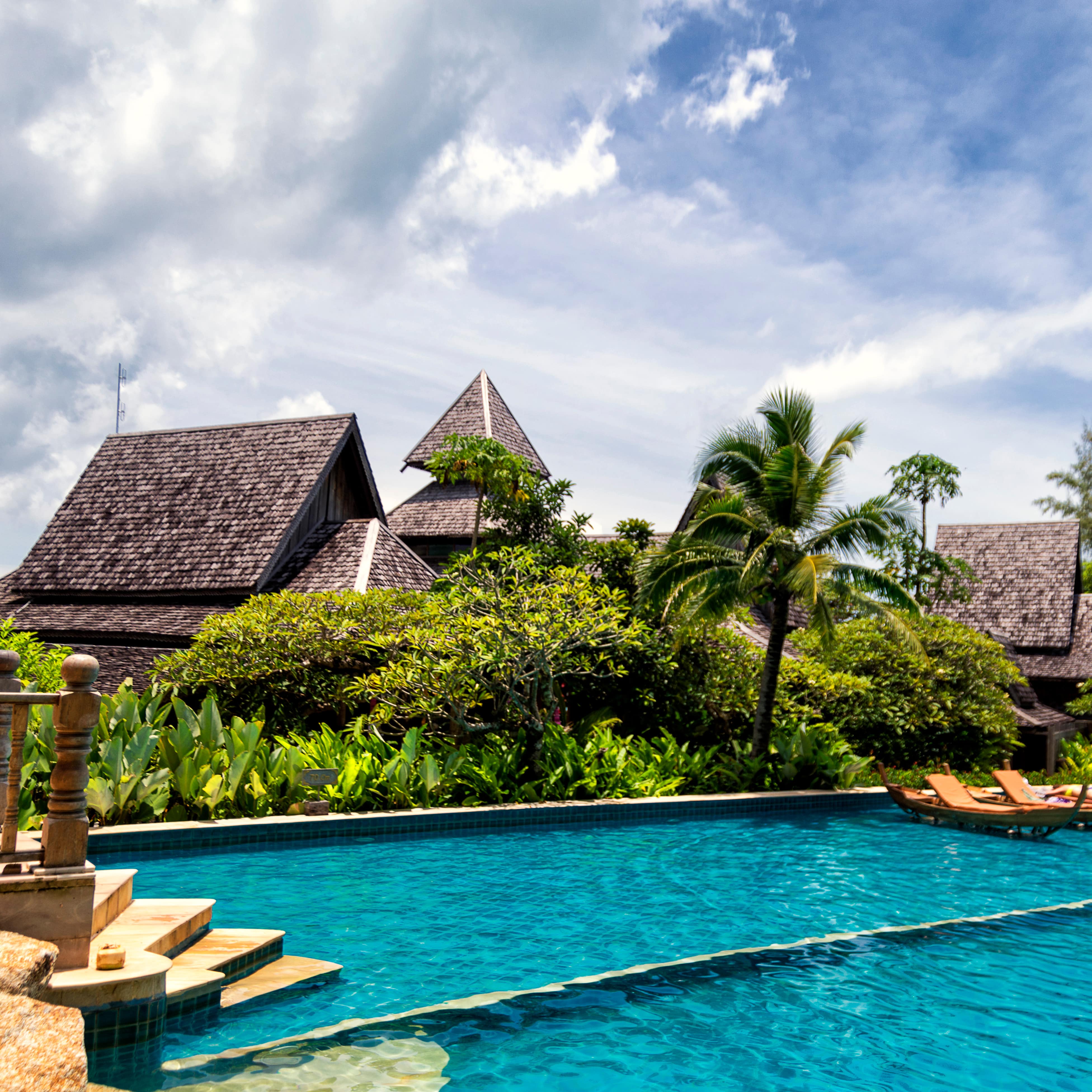 Pool with floating lounges against a backdrop of a Thai villa, palm trees, tropical plants and a dramatic sky