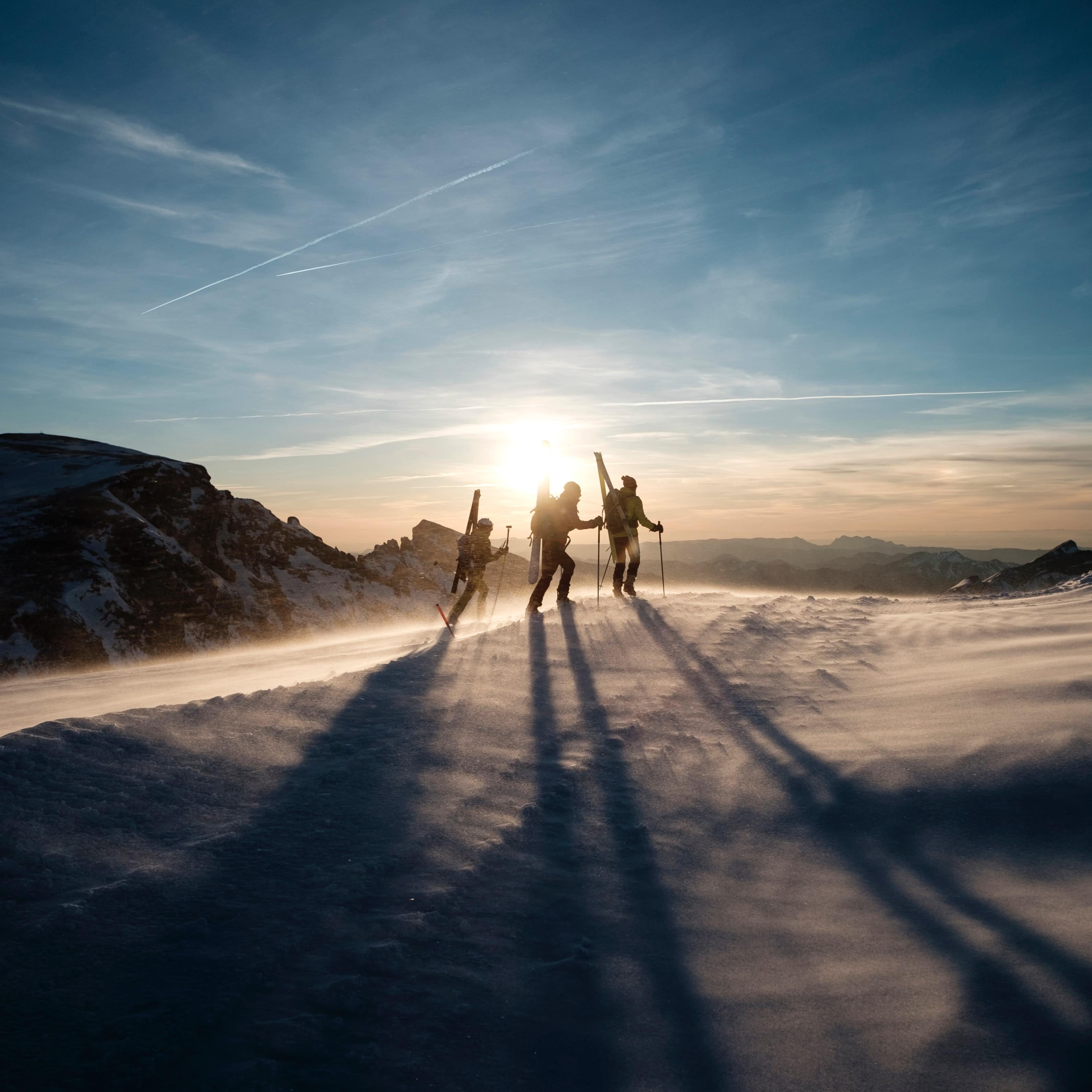 Silhouettes of three skiers walking up a snowy mountainside with the sun in the background