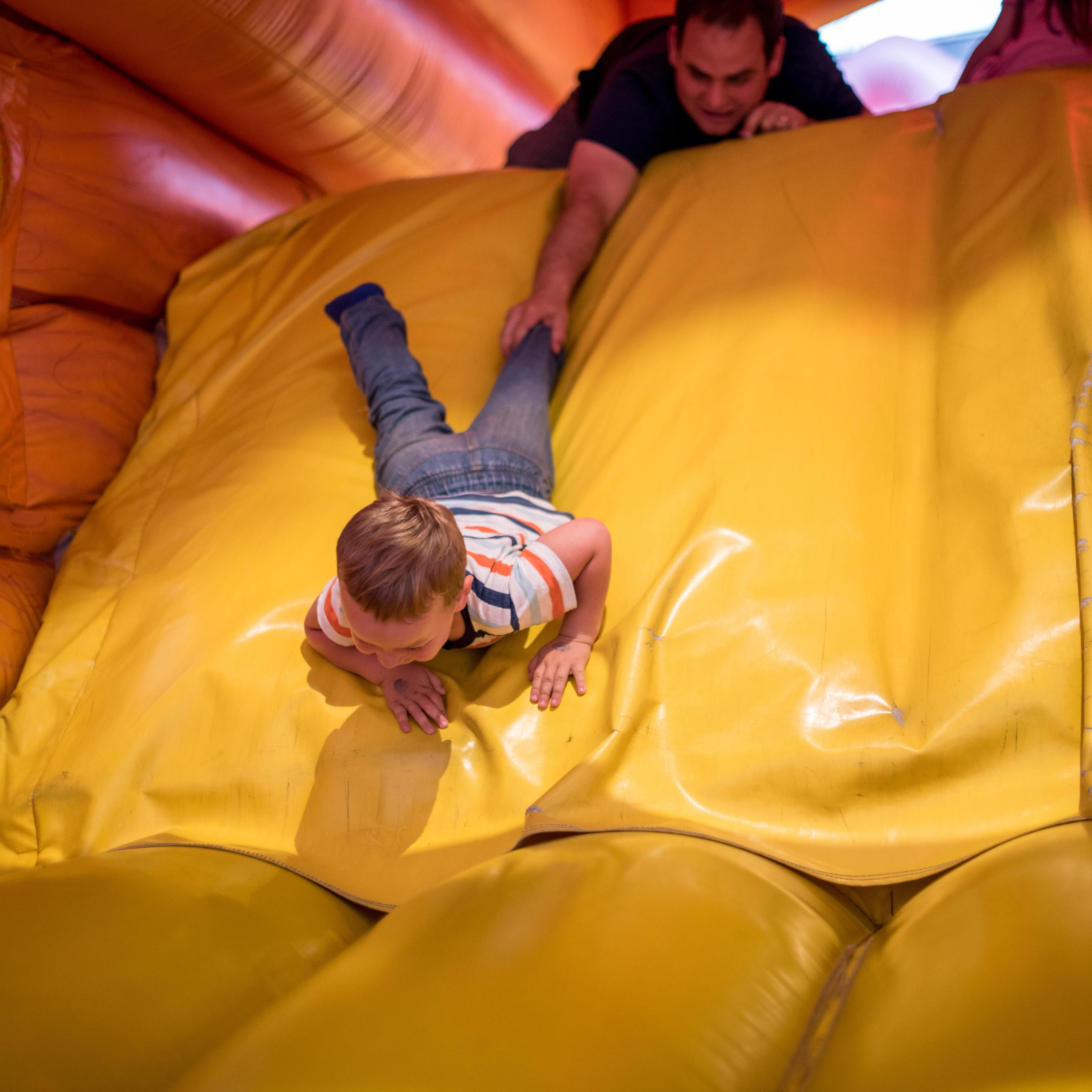 A boy sliding down an inflatable slide while his dad grabs his leg in an indoor playground