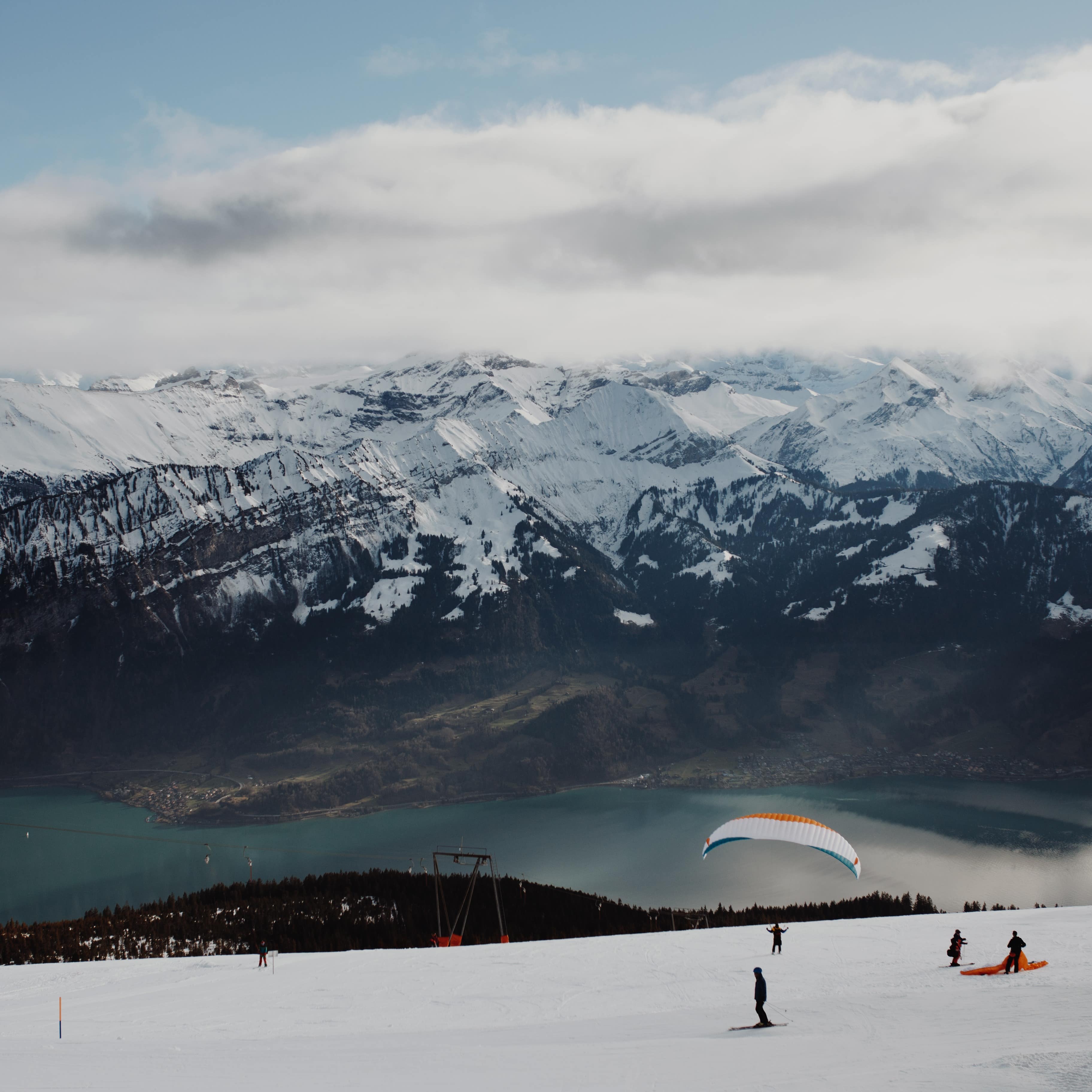 Skiers paragliding onto the slopes towards a beautiful lake surrounded by snowy mountain range.