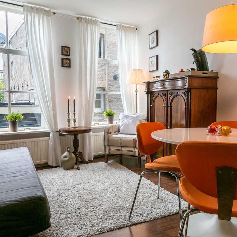 Vintage living room with hardwood floors, grey sofa, beige shag rug, round dining set, tall windows with white curtains.