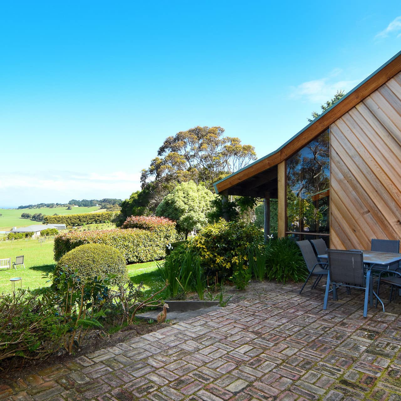 Bricked patio and outdoor dining table outside 4-bedroom stained-wood farmhouse set in lovely farmland with shrubs and trees