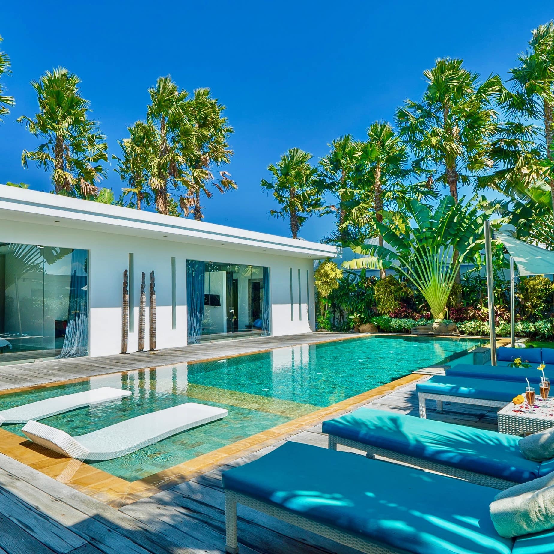 Resort pool with blue lounge chairs and white pool floats, surrounded by palm trees and lush vegetation.