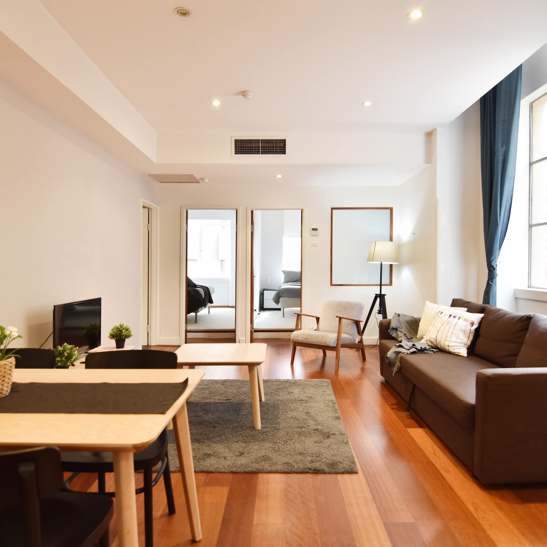 Open living and dining room with hardwood floors, white walls, brown and beige furnishings, large window with blue curtains.