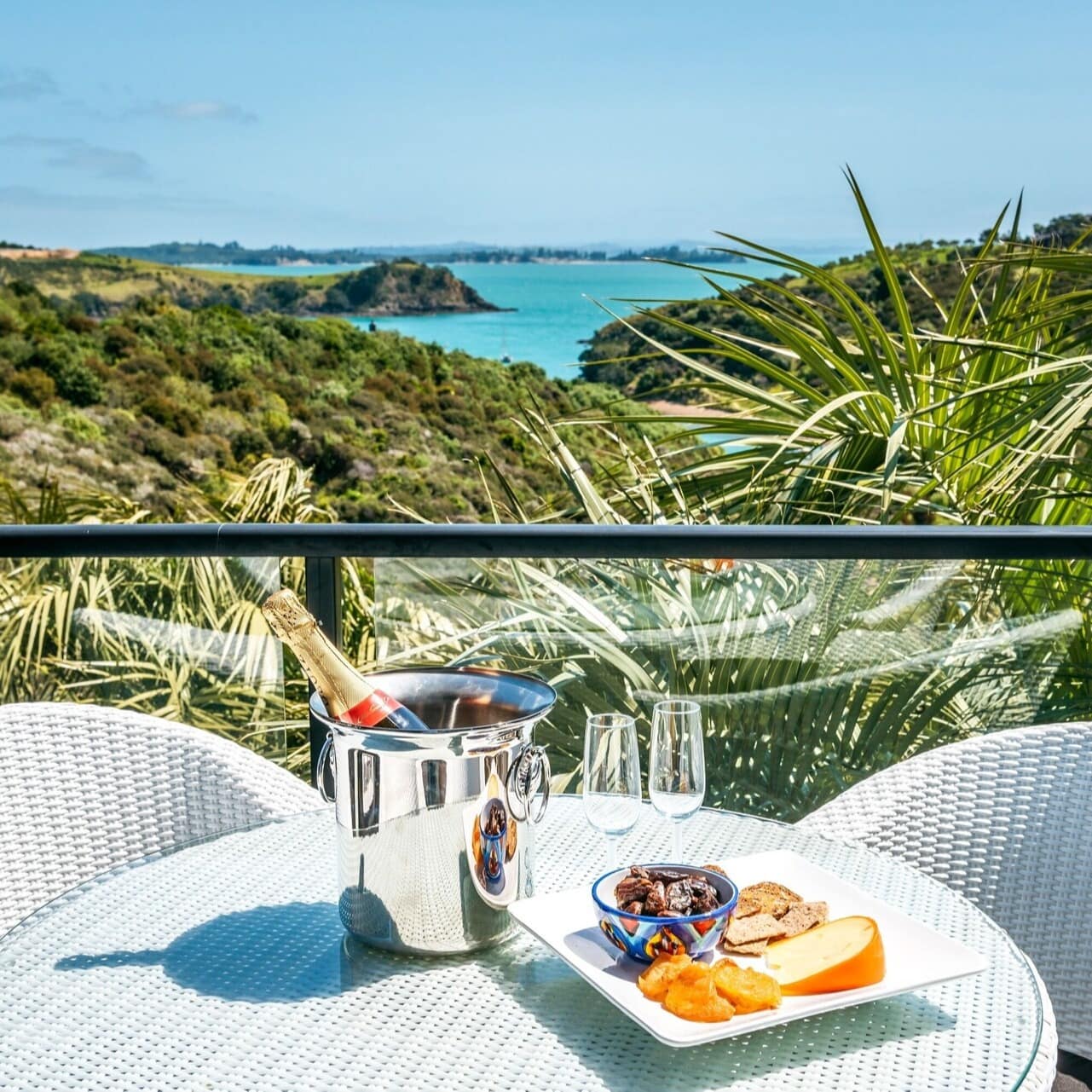A set table with chairs on the back deck of a holiday home overlooking green hills and water in the background