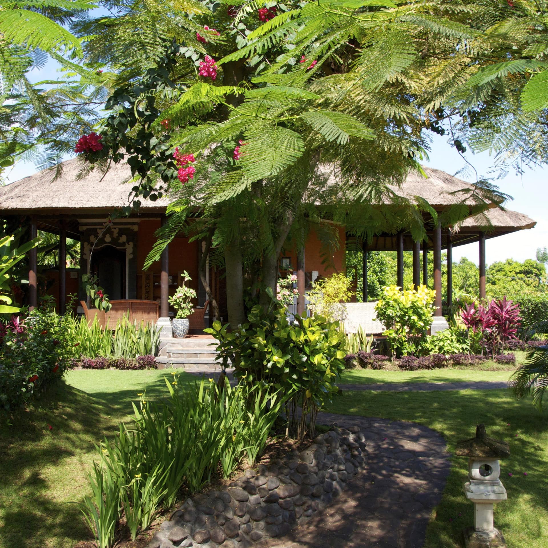 Sunny view of a villa with a straw roof and lush lawn with tropical plants.