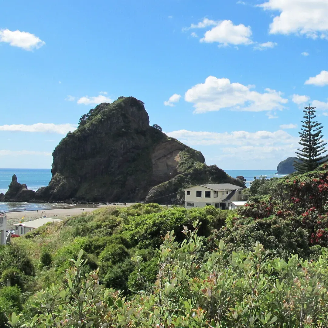 Oceanfront beach houses and cliffs behind luscious green hills under a blue sky