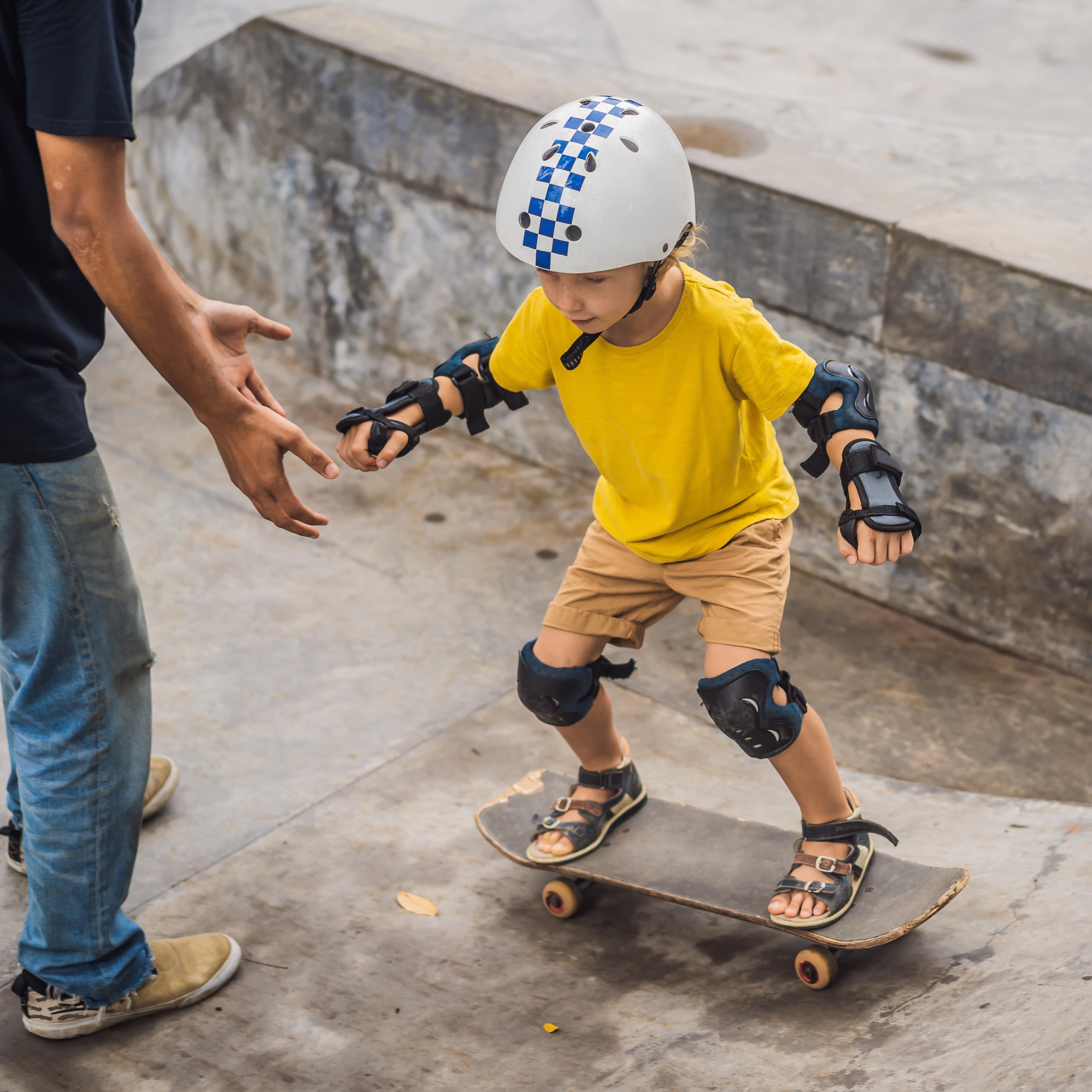 Young boy learns to skateboard in a skate park with man holding hands out for assistance