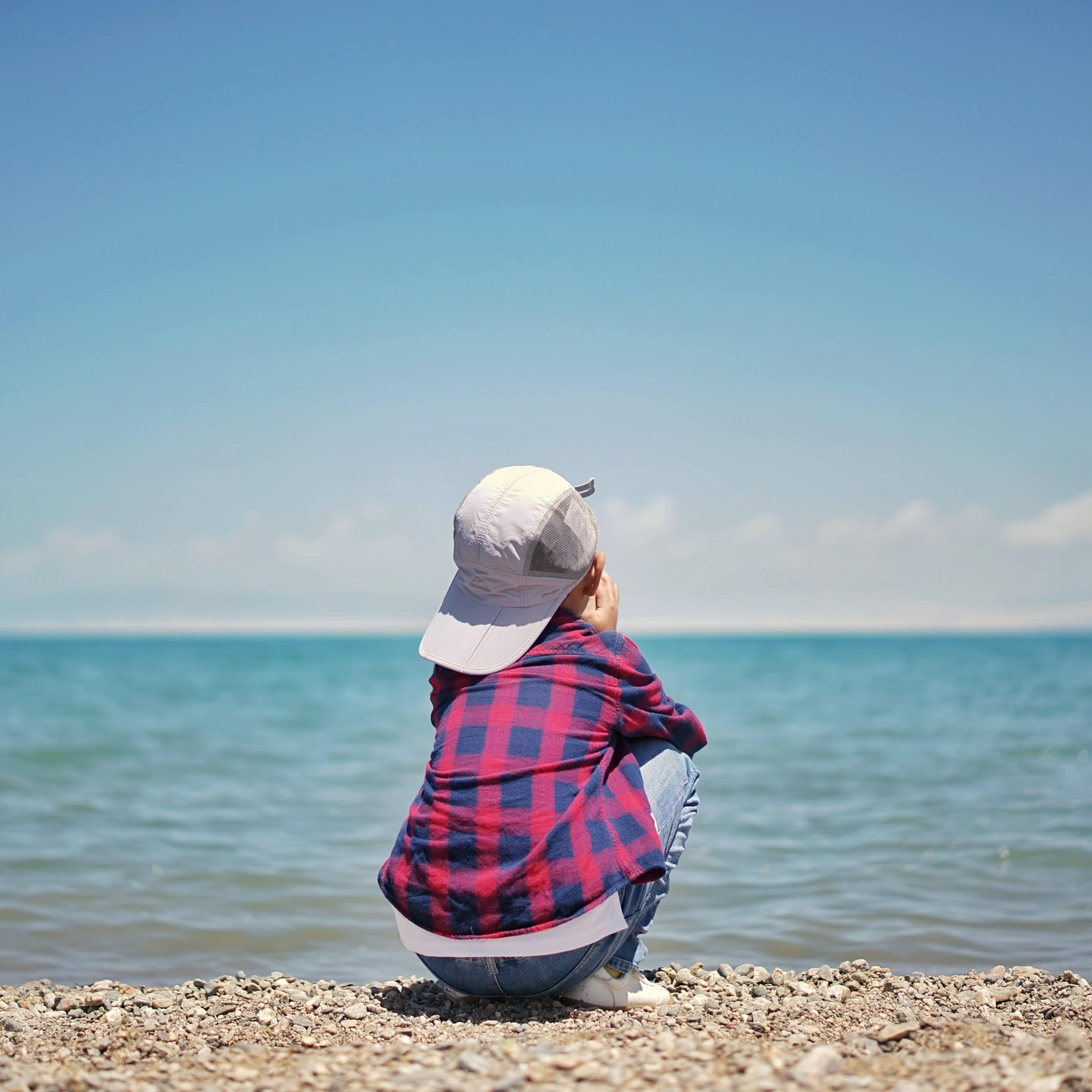 Child in a plaid shirt and hat crouching and looking out on the ocean from behind on a rocky shore