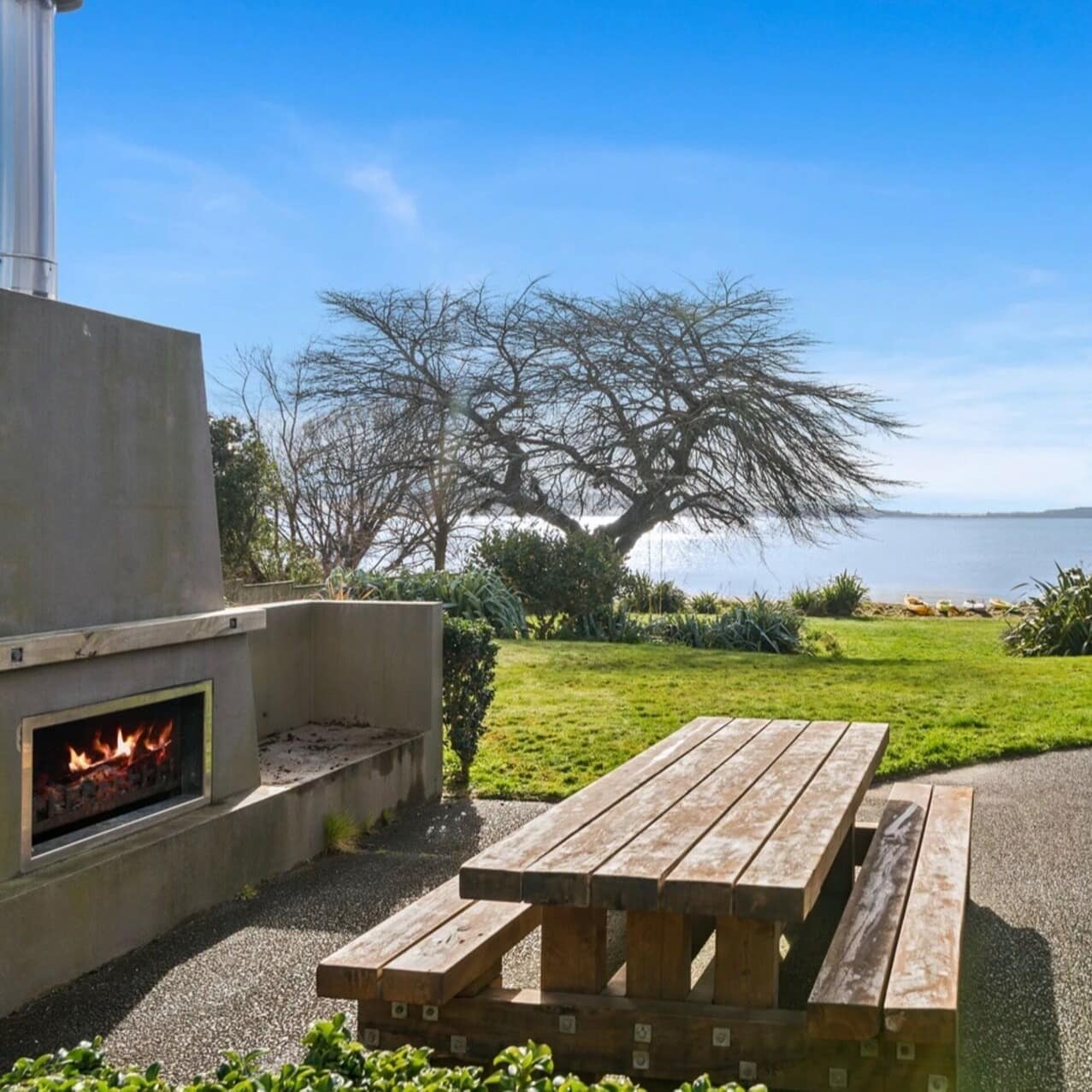 The back patio of a family home with a wooden picnic table and fireplace looking out on the lake with trees and kayaks on the shore