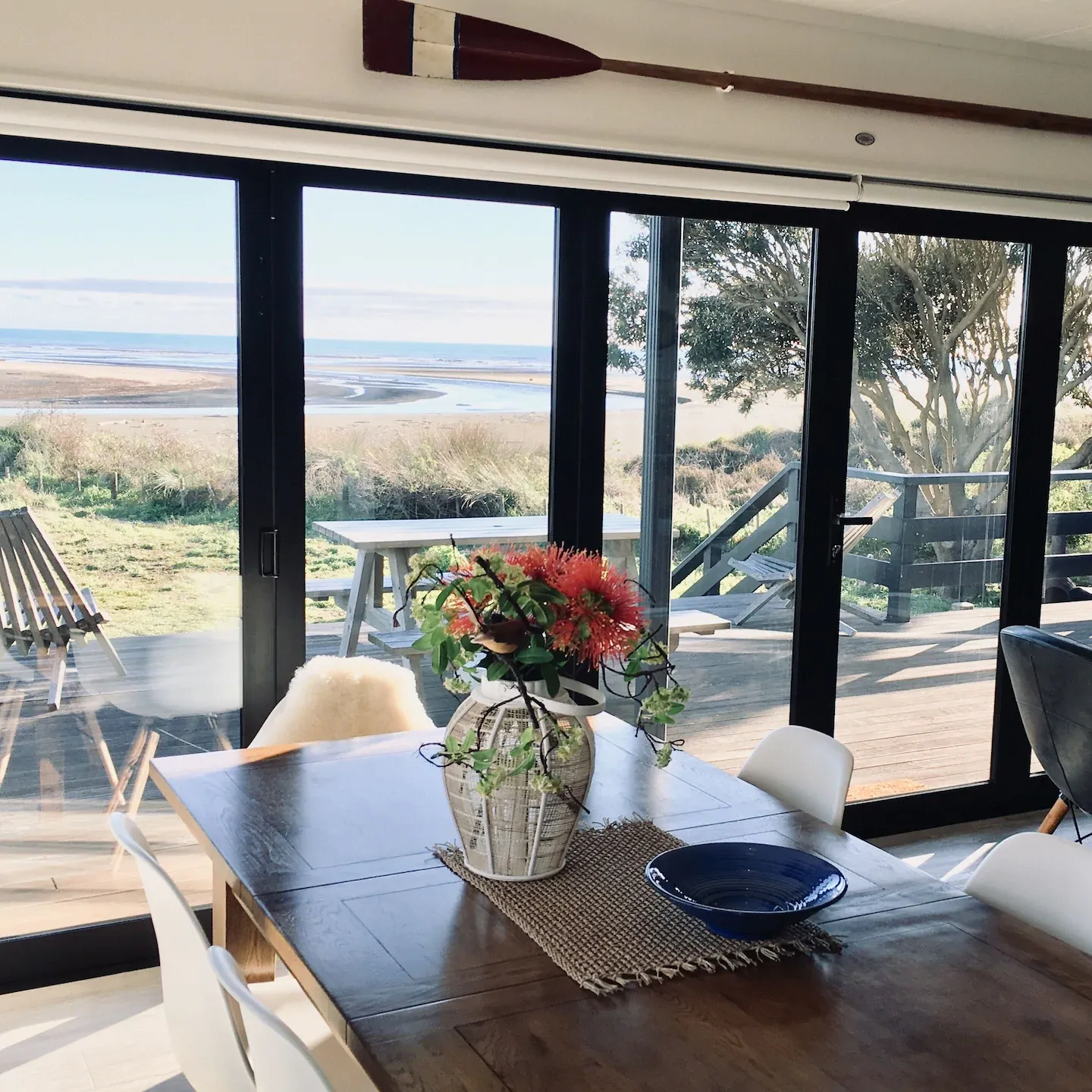 View of the beach and vegetation from the inside of a holiday home, with a table set with a flowerpot in the foreground