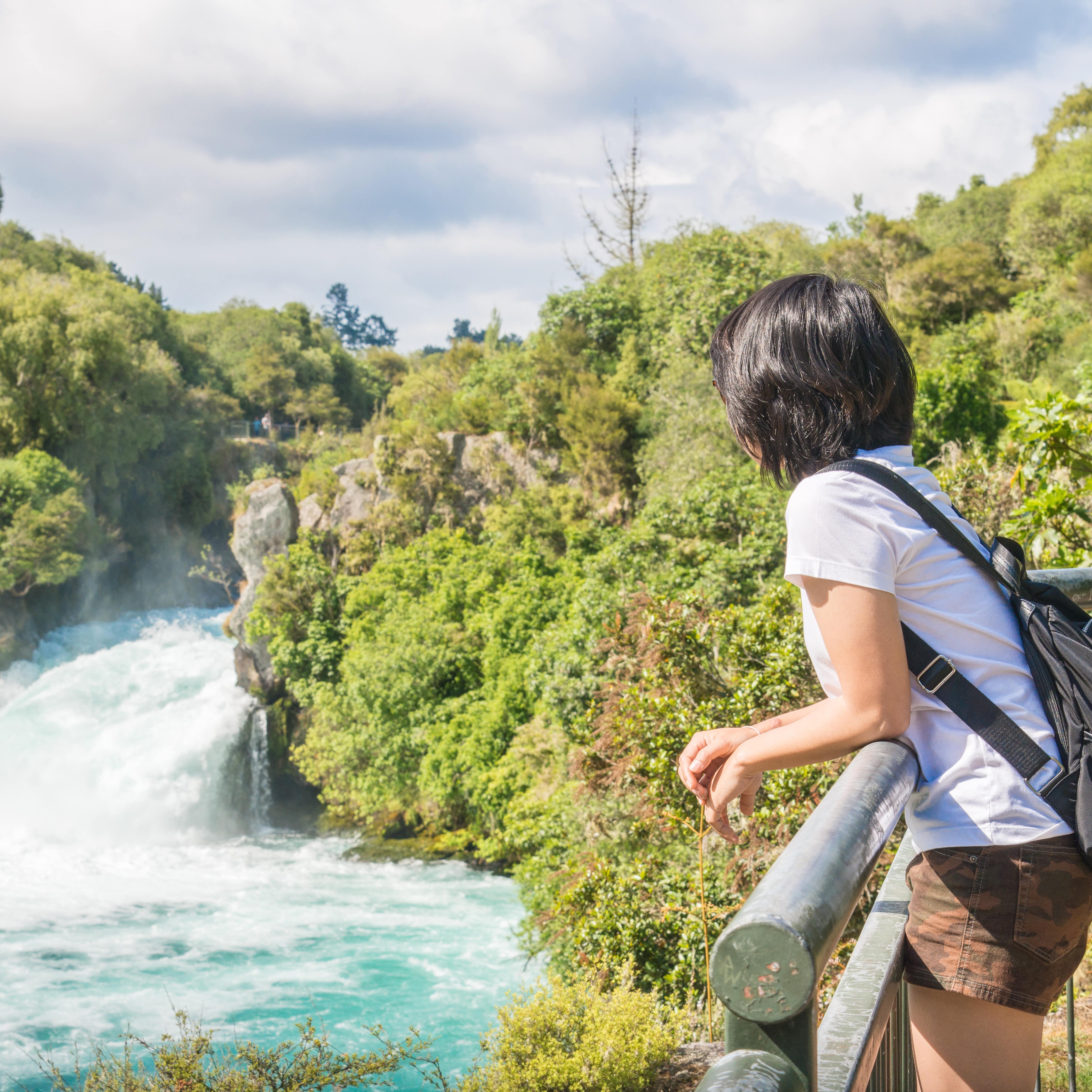Asian woman viewing aqua green Huka Falls, Waikato River, Taupo District, New Zealand with people on cliff above looking at rushing waters