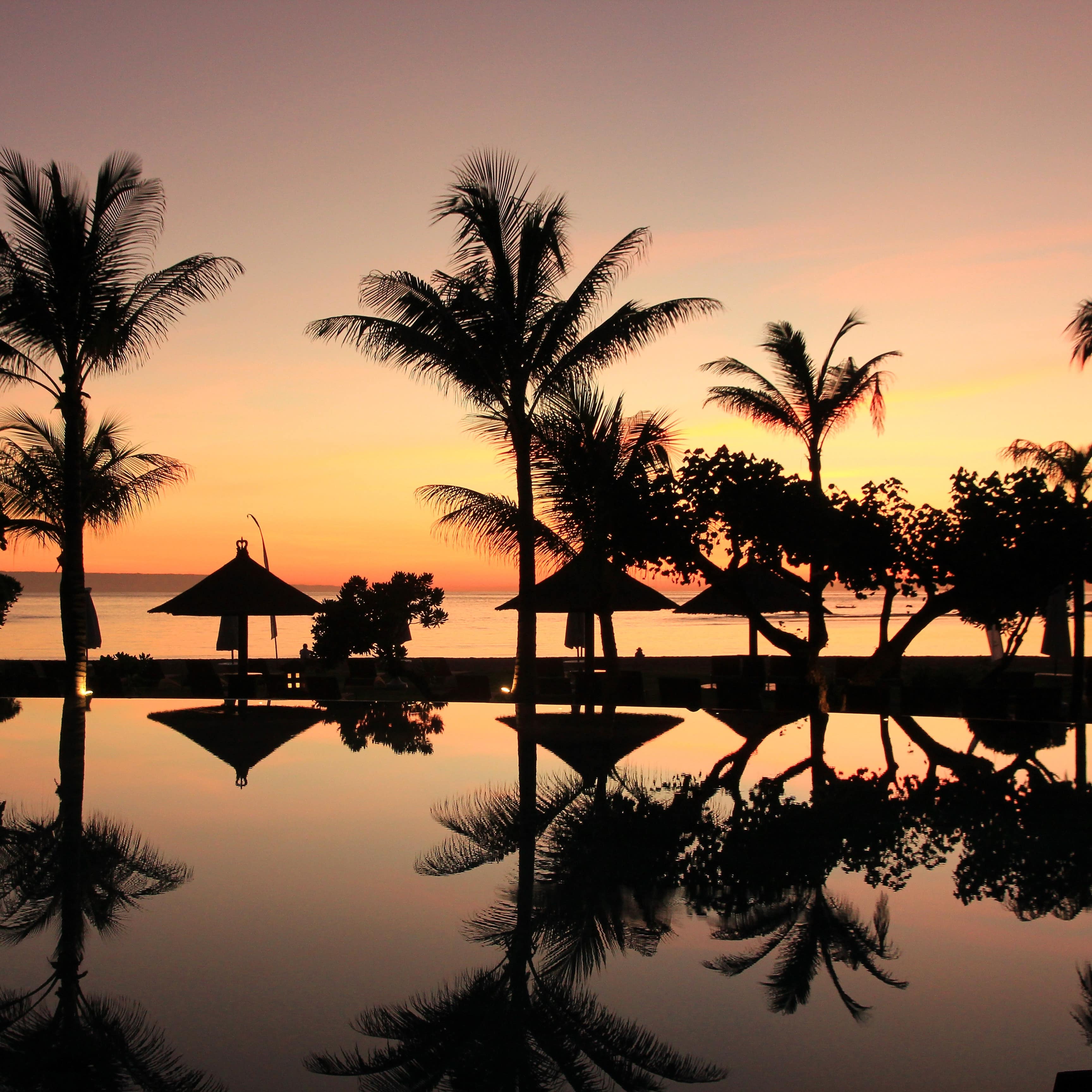 Beachside pool at sunset with dark reflections of palm trees and beach umbrellas and the Indian Ocean beyond