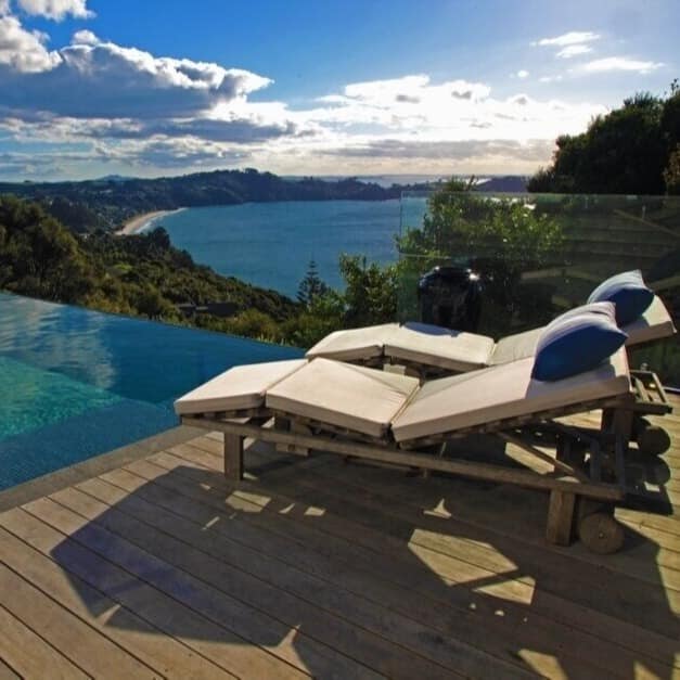 A wooden deck with a pool and lounge chairs overlooking the bright blue ocean from a clifftop home