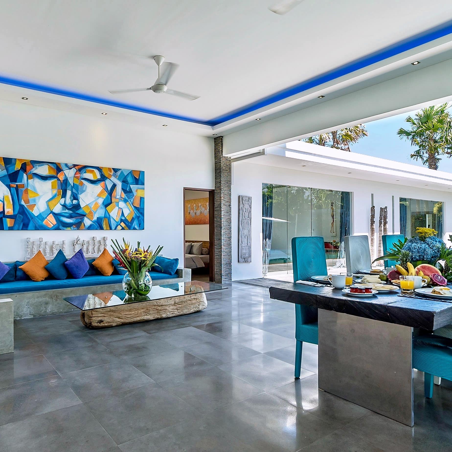 An open living and dining room with grey tile floors, wooden dining table, blue chairs, blue sectional, glass coffee table.
