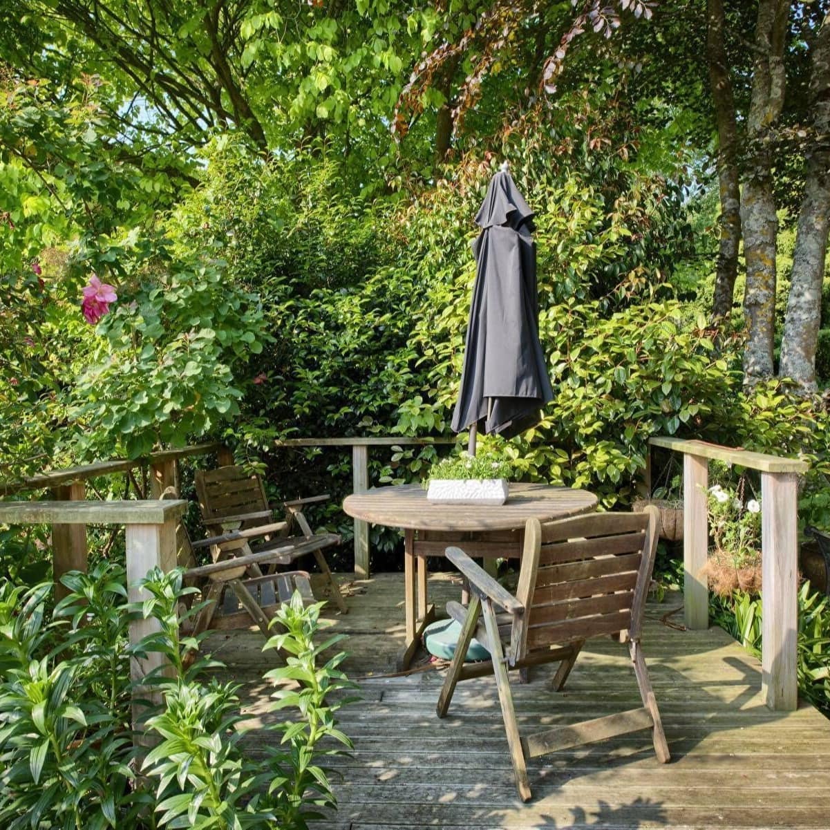 Wooden deck of a cottage with chairs around a small shaded table, surrounded by lush trees and bushes