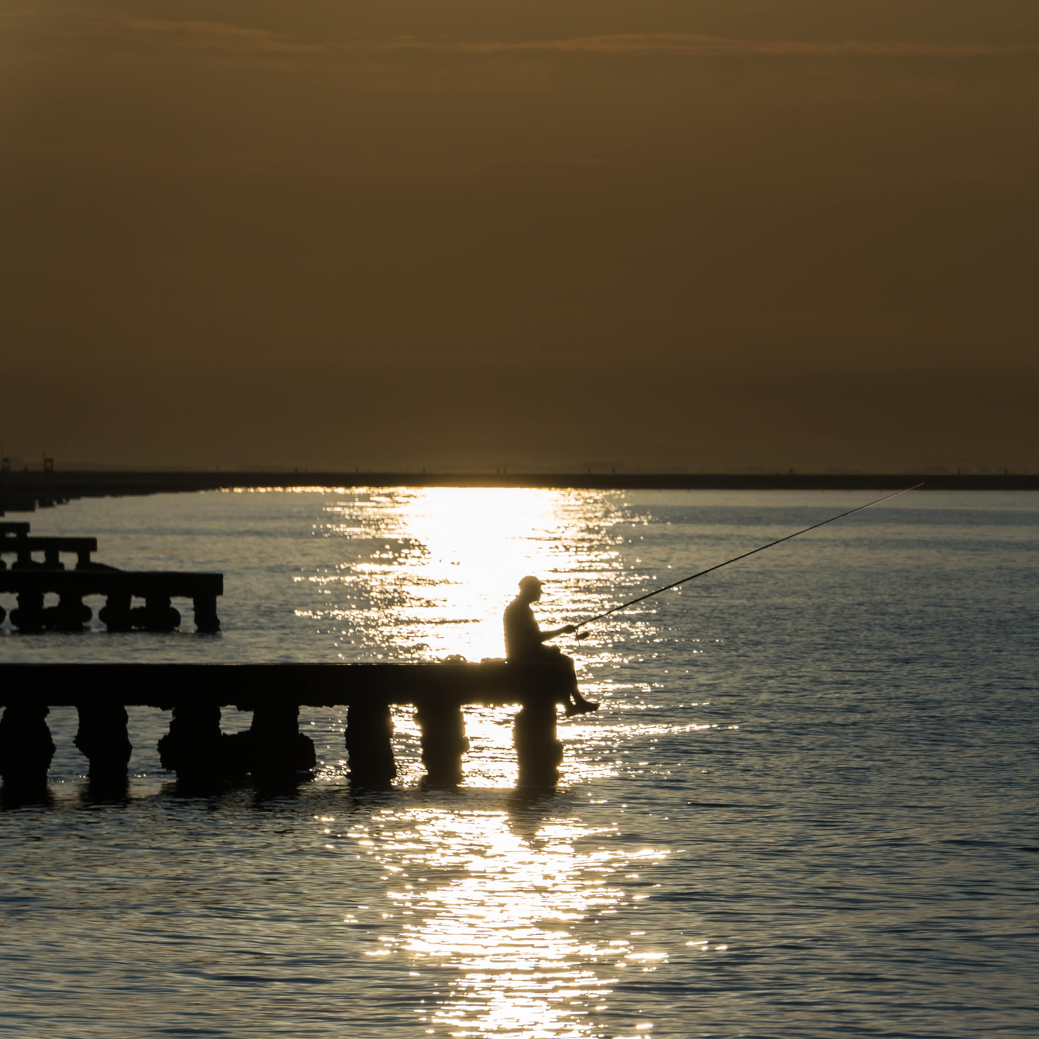 A man sitting on the dock fishing as the sunsets over the ocean.