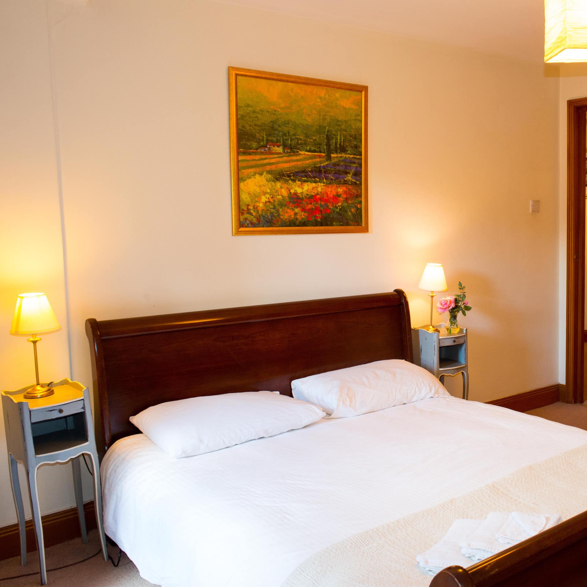 A warm bedroom with a wood-framed king size bed, white bedding, vintage bedside tables, tall wooden wardrobes.