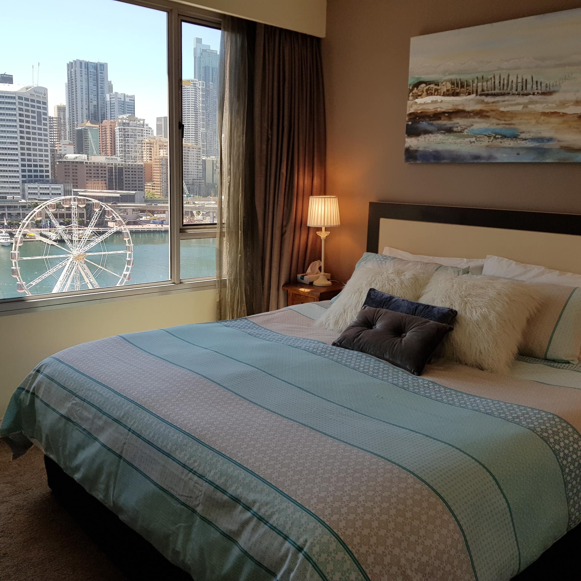 Bedroom with a king size bed, teal and white bedding, and wide windows with views of the city skyline, and harbour.