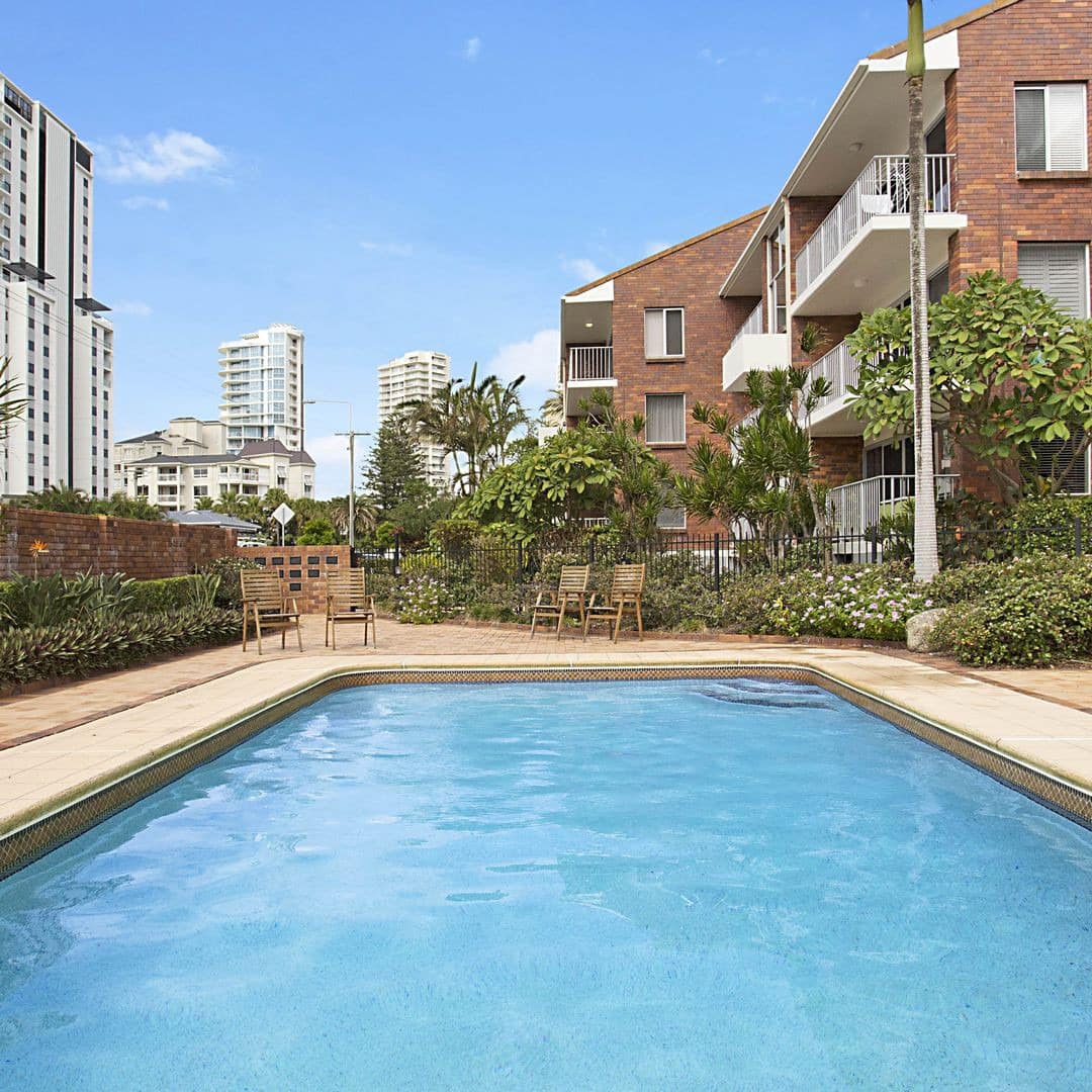 View of an inground pool with beige tile pool deck next to an apartment building.