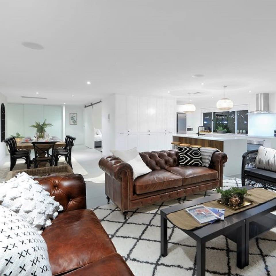 Modern open living room and kitchen with brown leather sofas, black rattan chairs, and a sleek white kitchen.