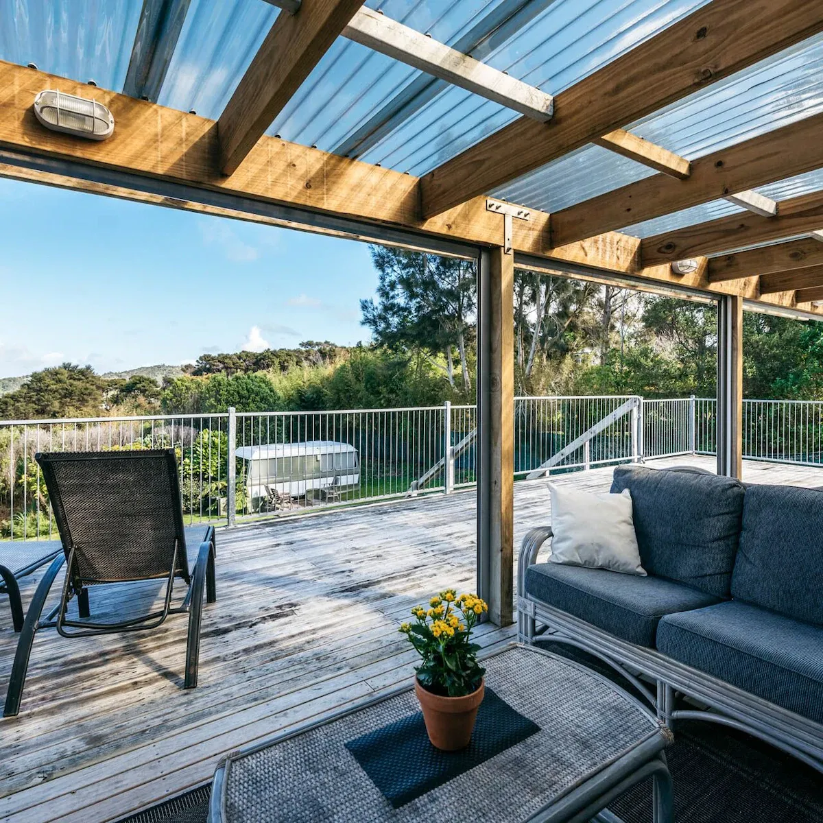 Back deck of a beach bach with chairs and a small coffee with a view of the ocean and surrounding nature in the background
