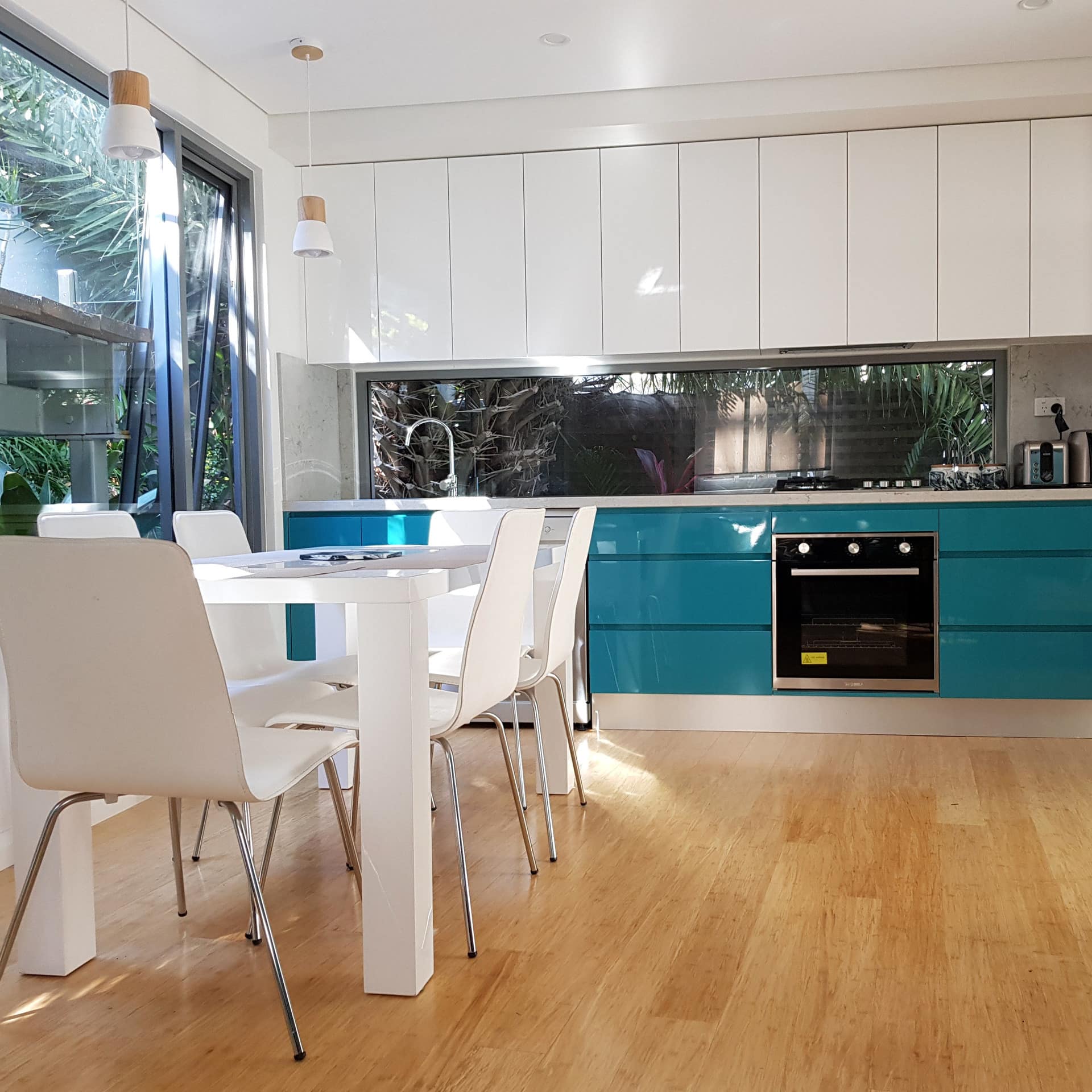 Modern, open kitchen and dining room with hardwood floors, white dining set, large windows with views of tropical plants.