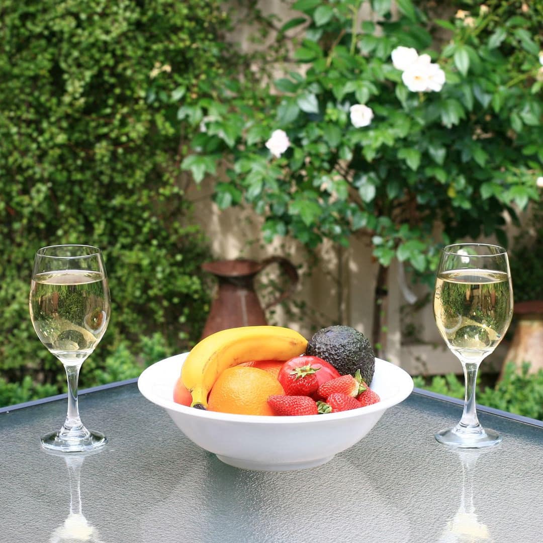 Wine and fruit sit on a glass table in front of lush greenery.