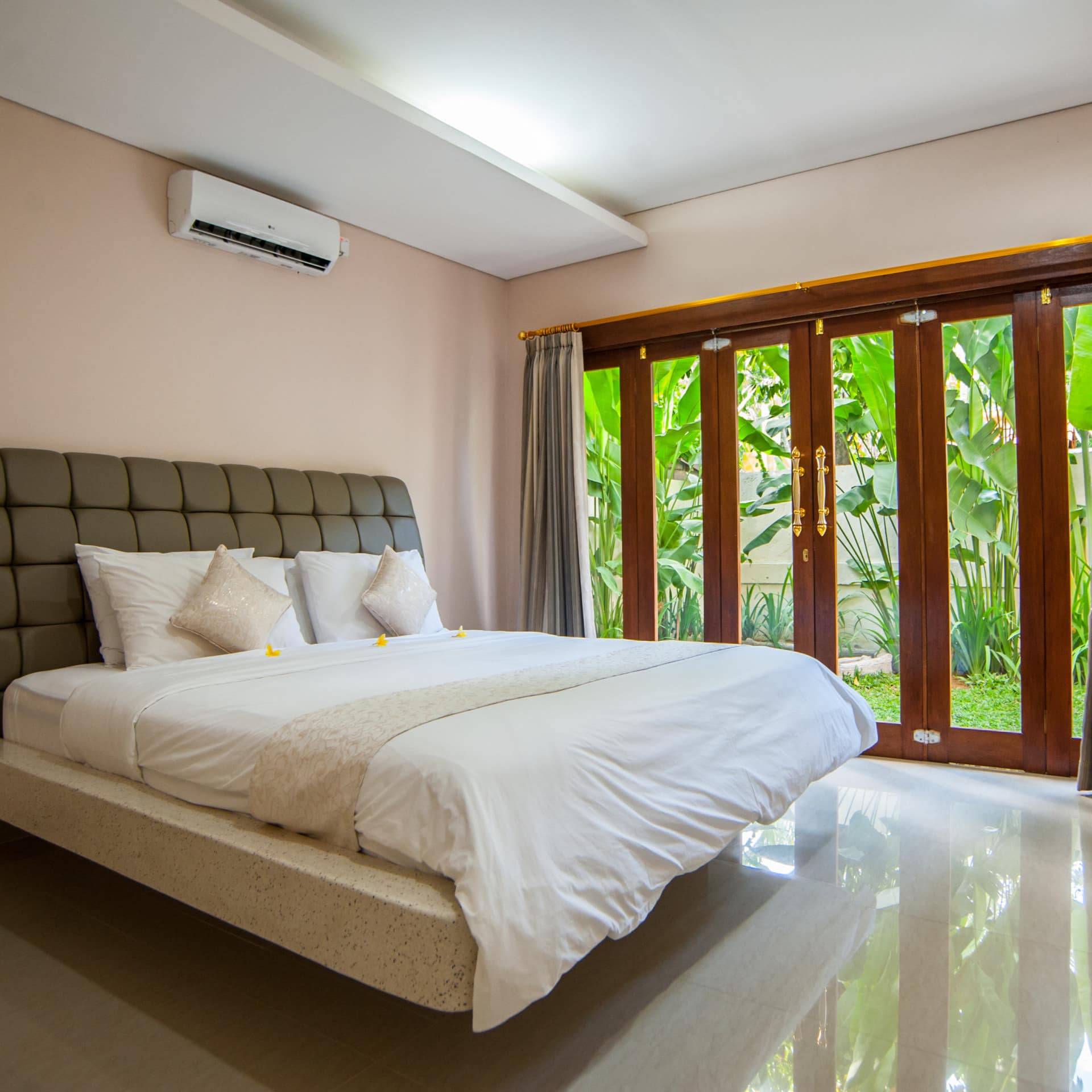 A bedroom with white stone floors, beige walls, a king size bed, white bedding, glass doors with views of tropical plants.
