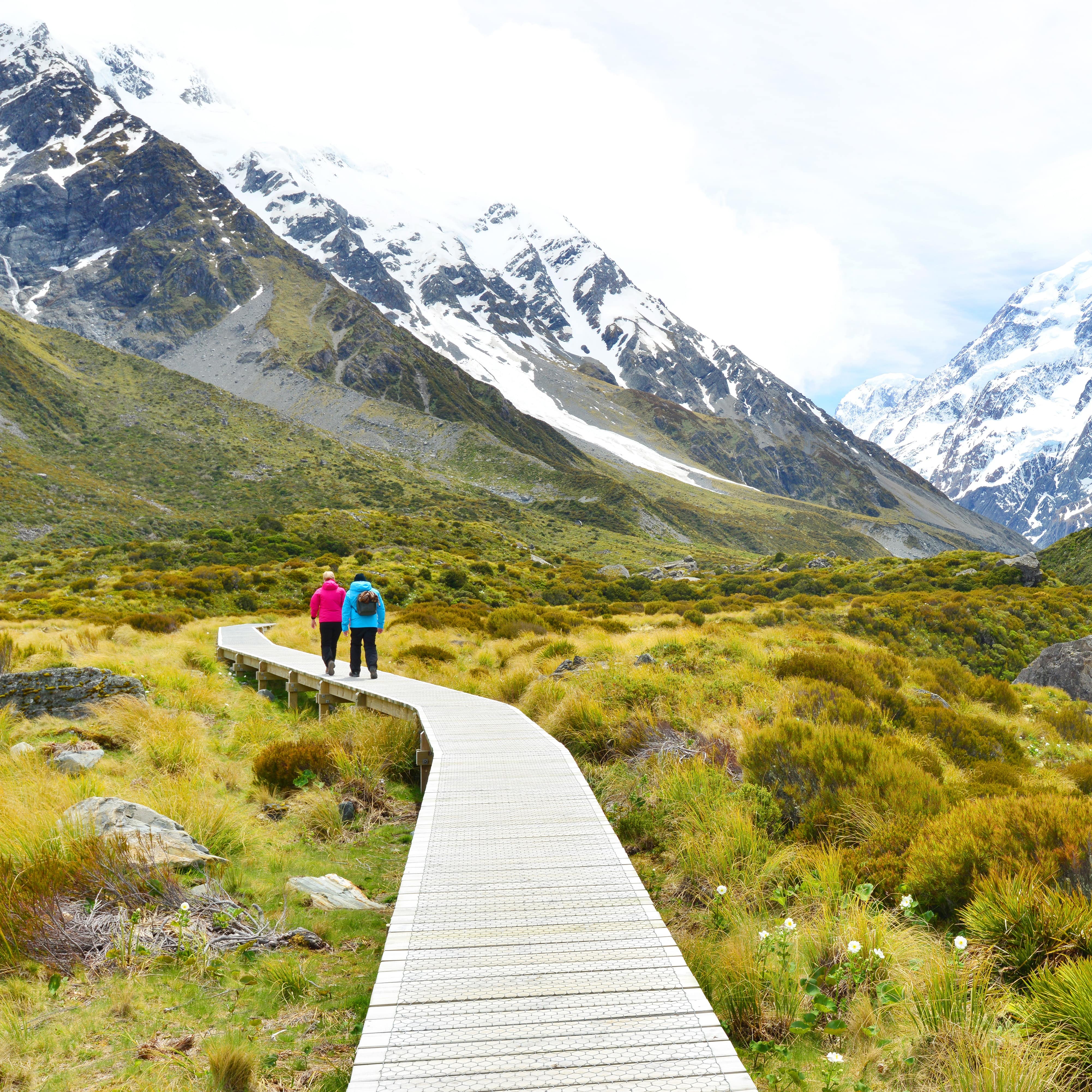 Two people walking on a bridge through the snow-capped Alps on a bright day