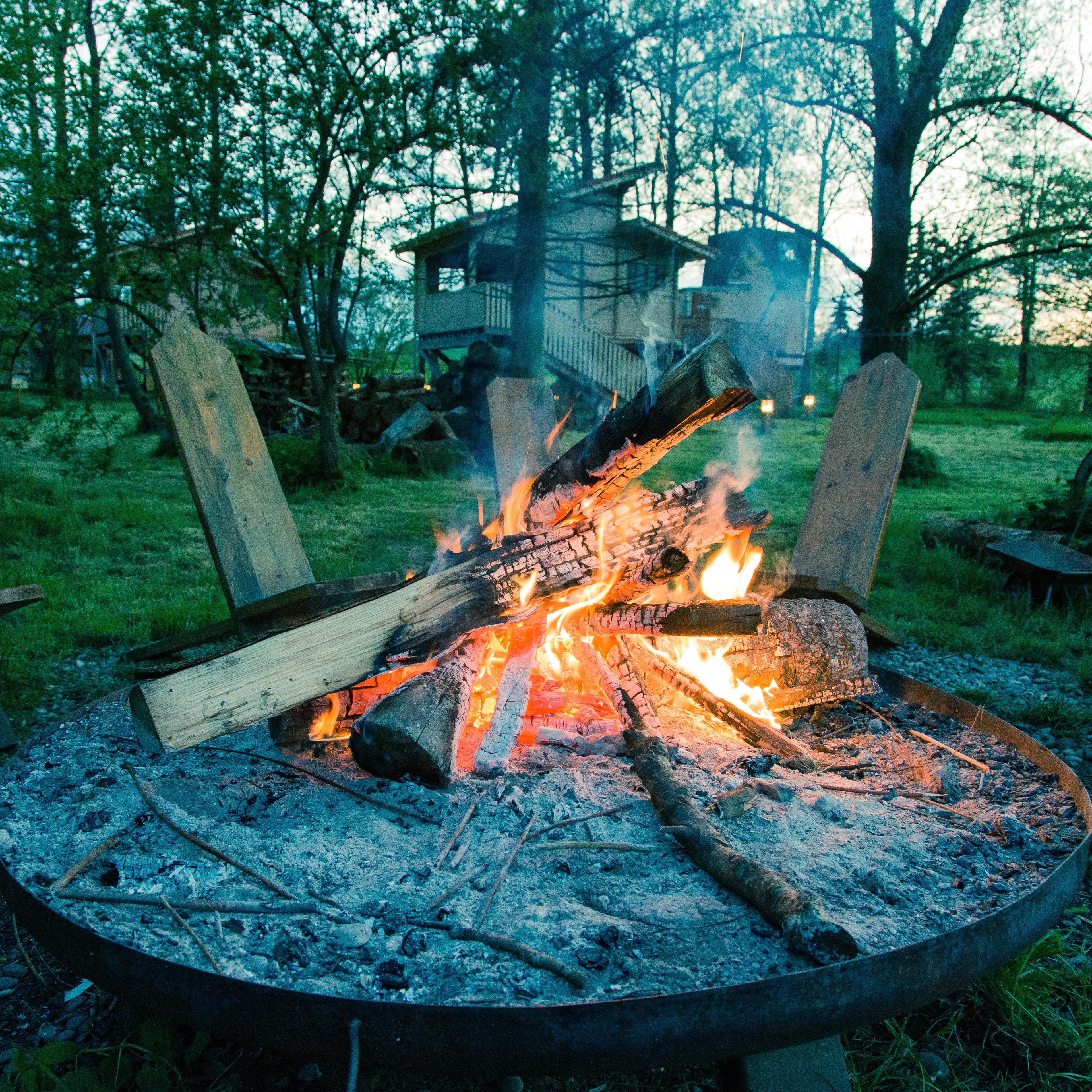 Fire pit with burning logs surrounded by Viking chairs with a camping pod situated in the forest