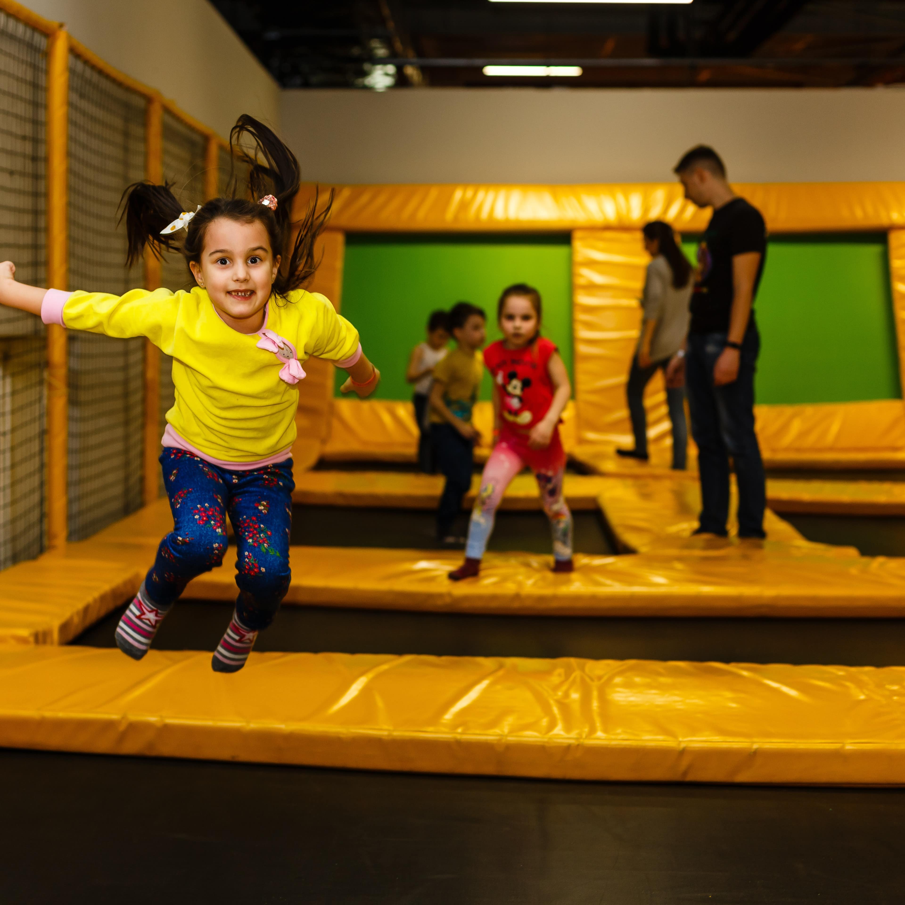 Little girl in yellow jumping on an indoor trampoline with more people in the background