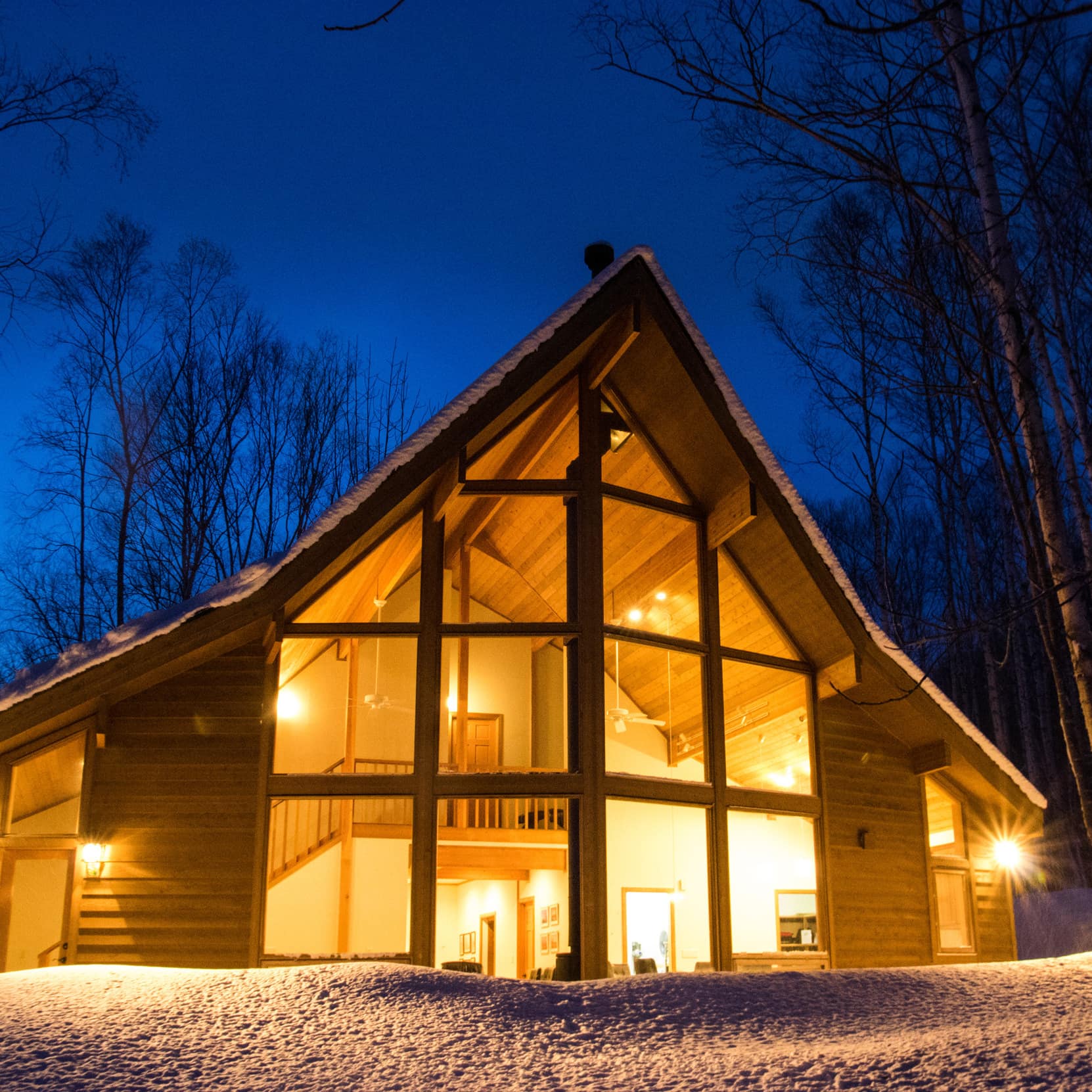 Nighttime view of an A-framed modern home with large windows, surrounded by forest and the ground covered by snow.