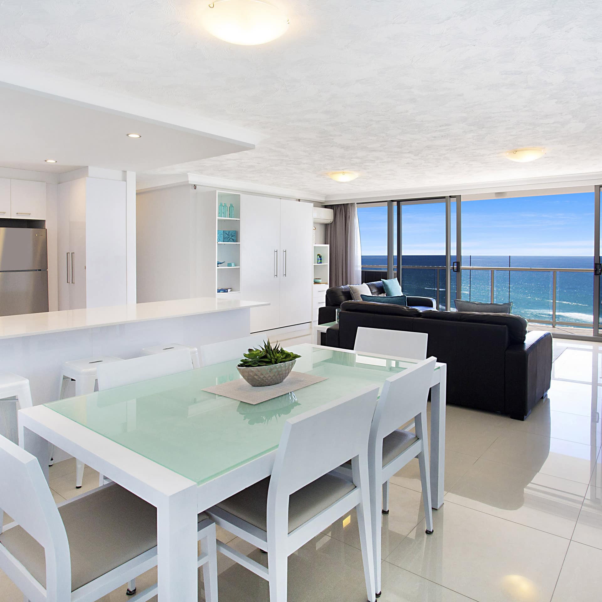 Modern apartment with white colour scheme including dining table, breakfast bar, kitchen, tile floor and grey leather sofa