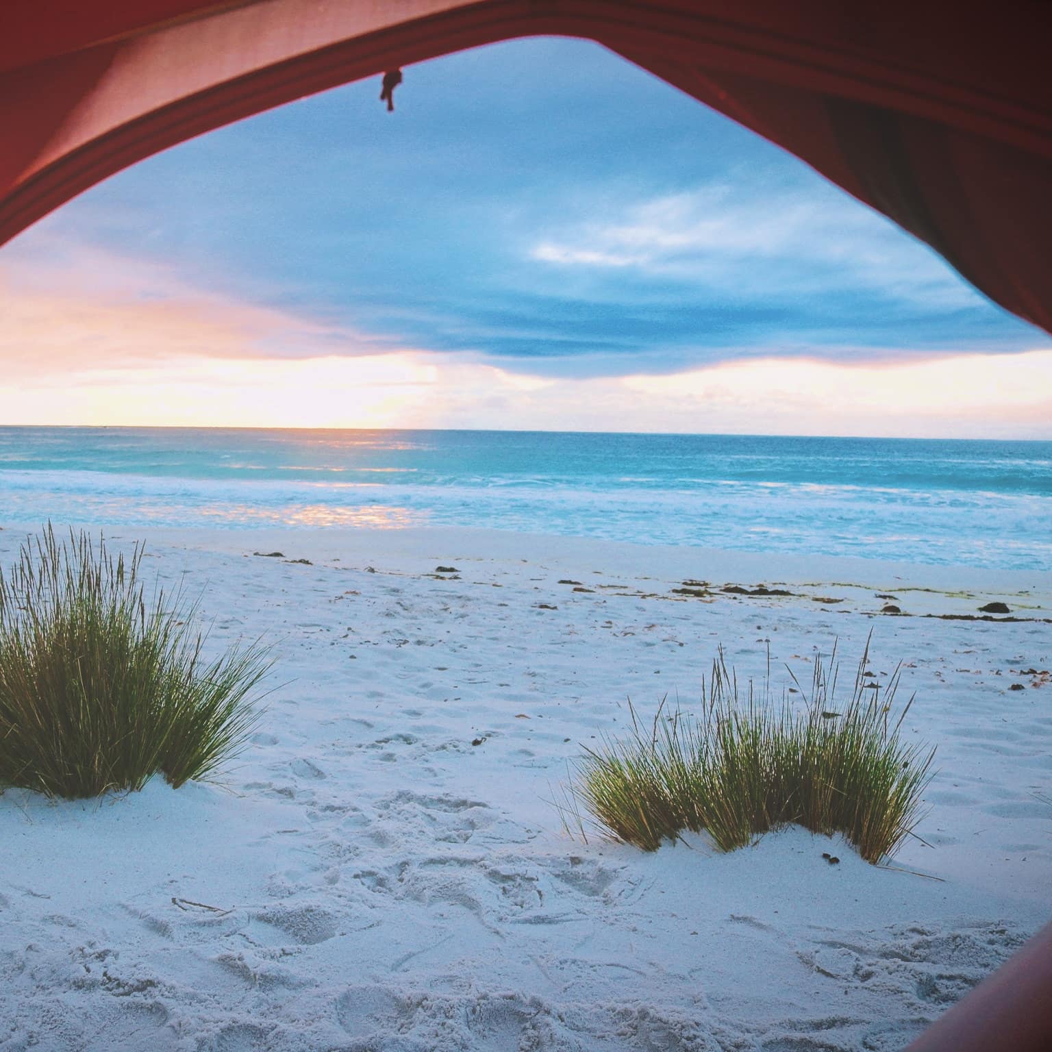A view from the opening of a red tent on an Aussie beach at sunrise with bright pink and blue skies and white sands