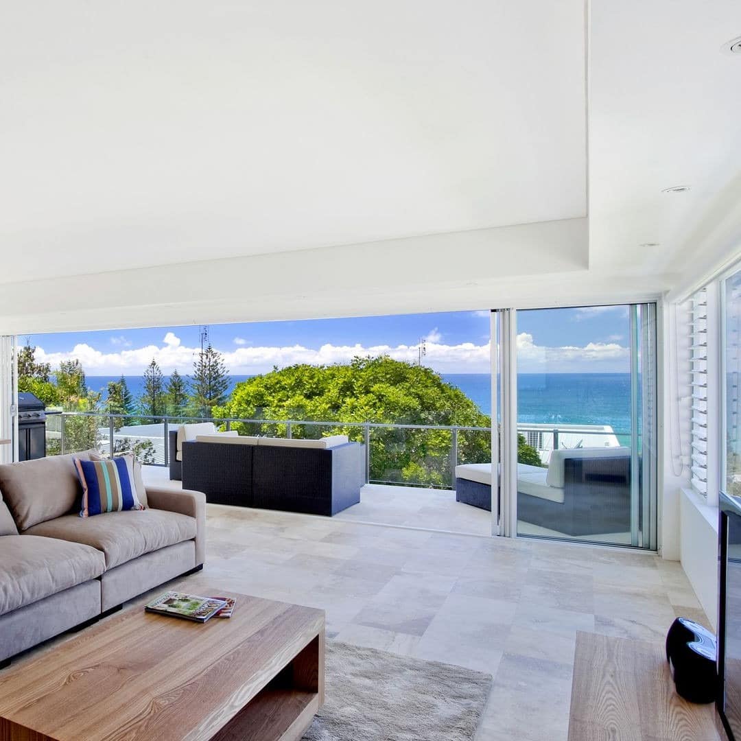 Open living room and kitchen with beige tile floors, a brown suede sofa, floor to ceiling windows with ocean views.