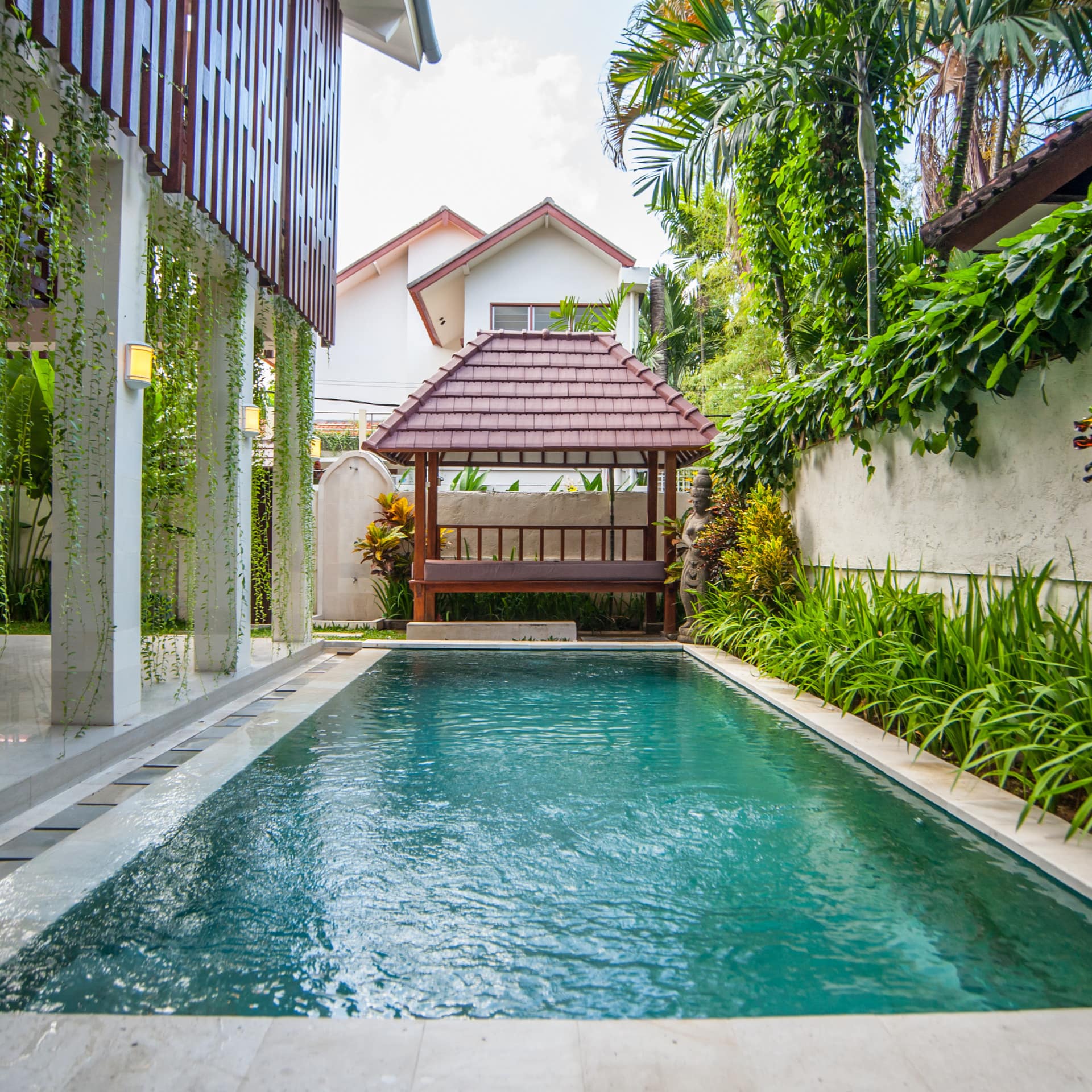 View of a pool with a covered patio space, a cabana, and surrounded by lush greenery.