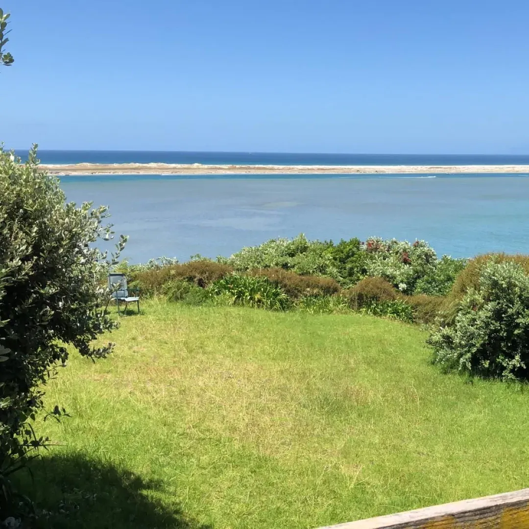 Close-up view of the beach from the wooden back deck of a beach bach with a small grassy backyard in the foreground
