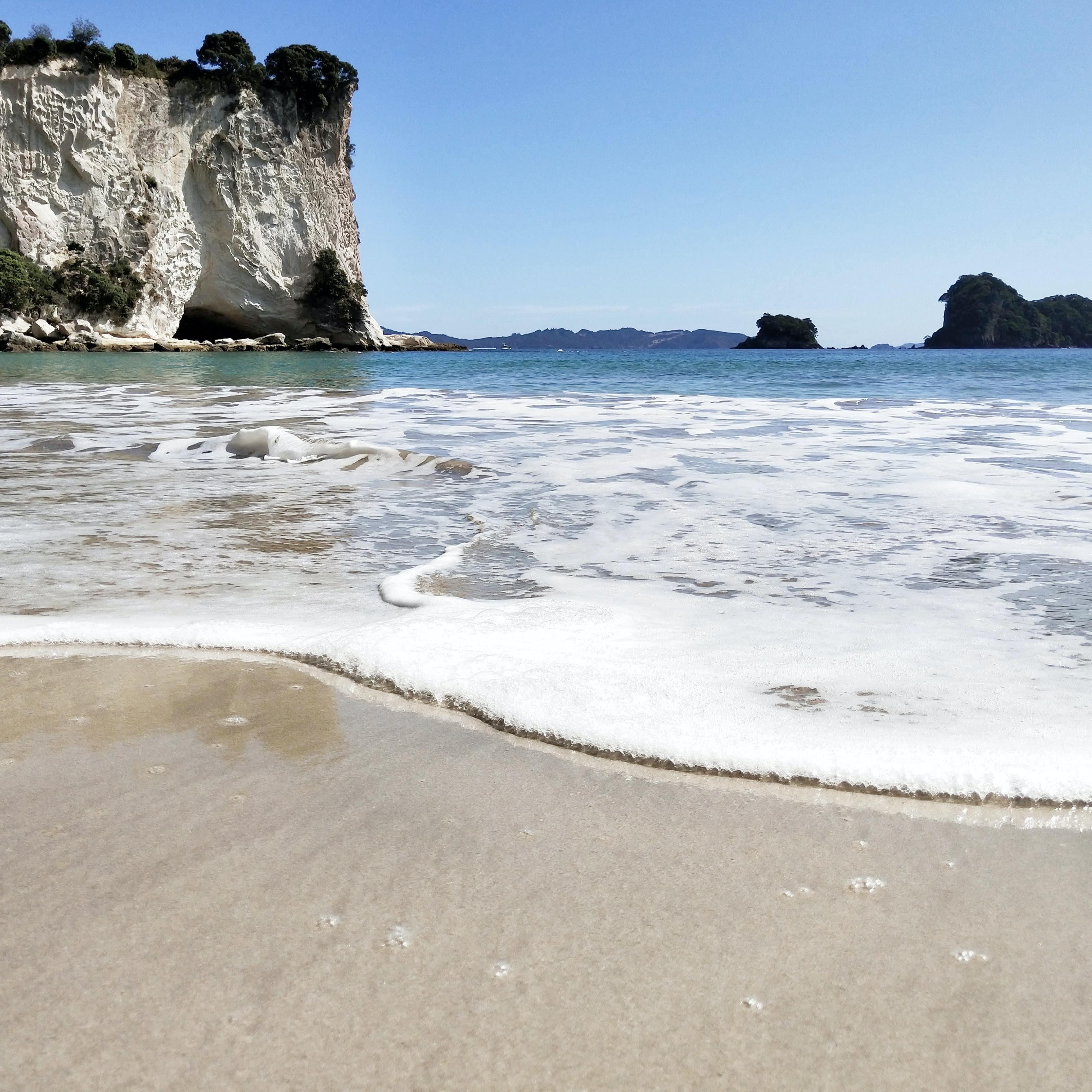Water crashing on a white, sandy shore with a large geological formation off the coast in Hahei, New Zealand